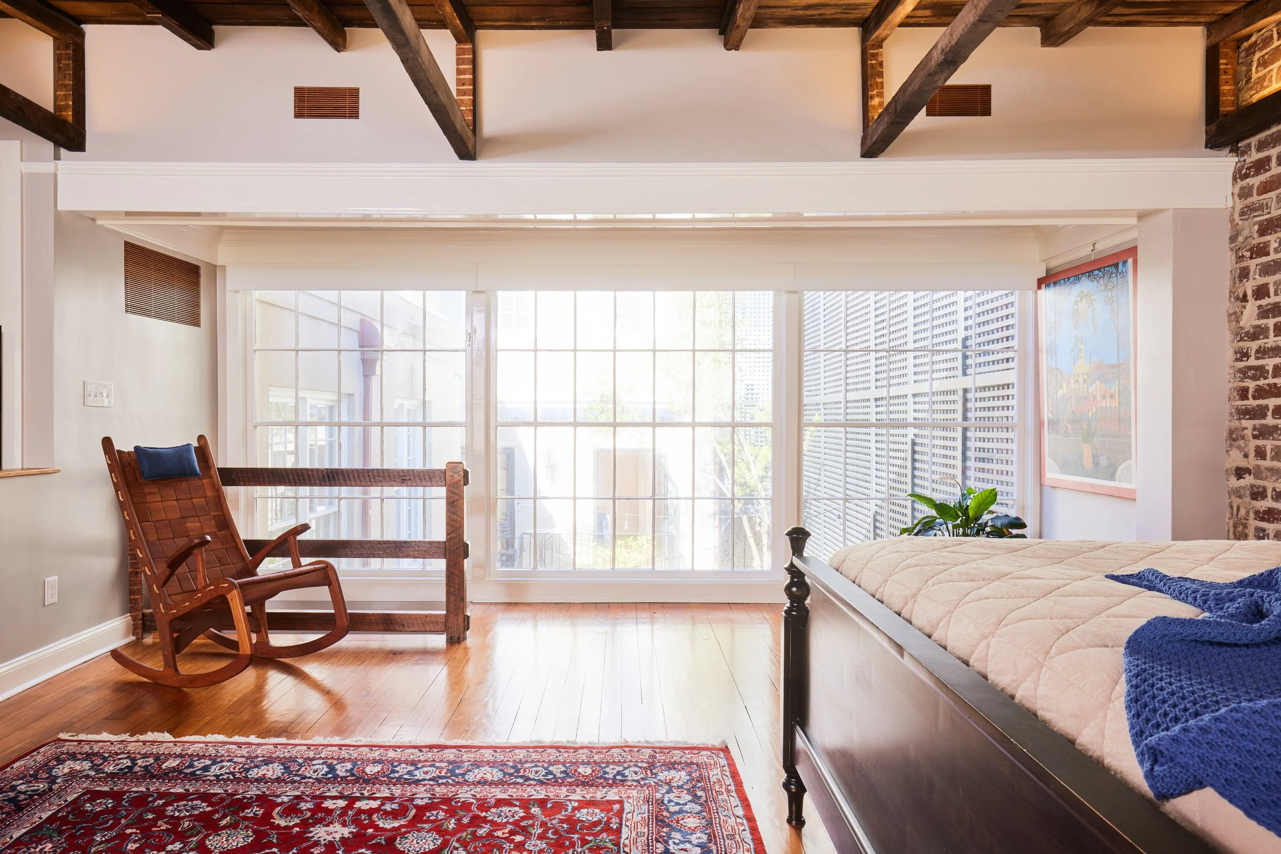 Bright bedroom with wooden ceiling beams, a large window at the back, a rocking chair, a bed with a beige quilt, a blue throw, a plant, a lamp, and a decorative painting on the wall.