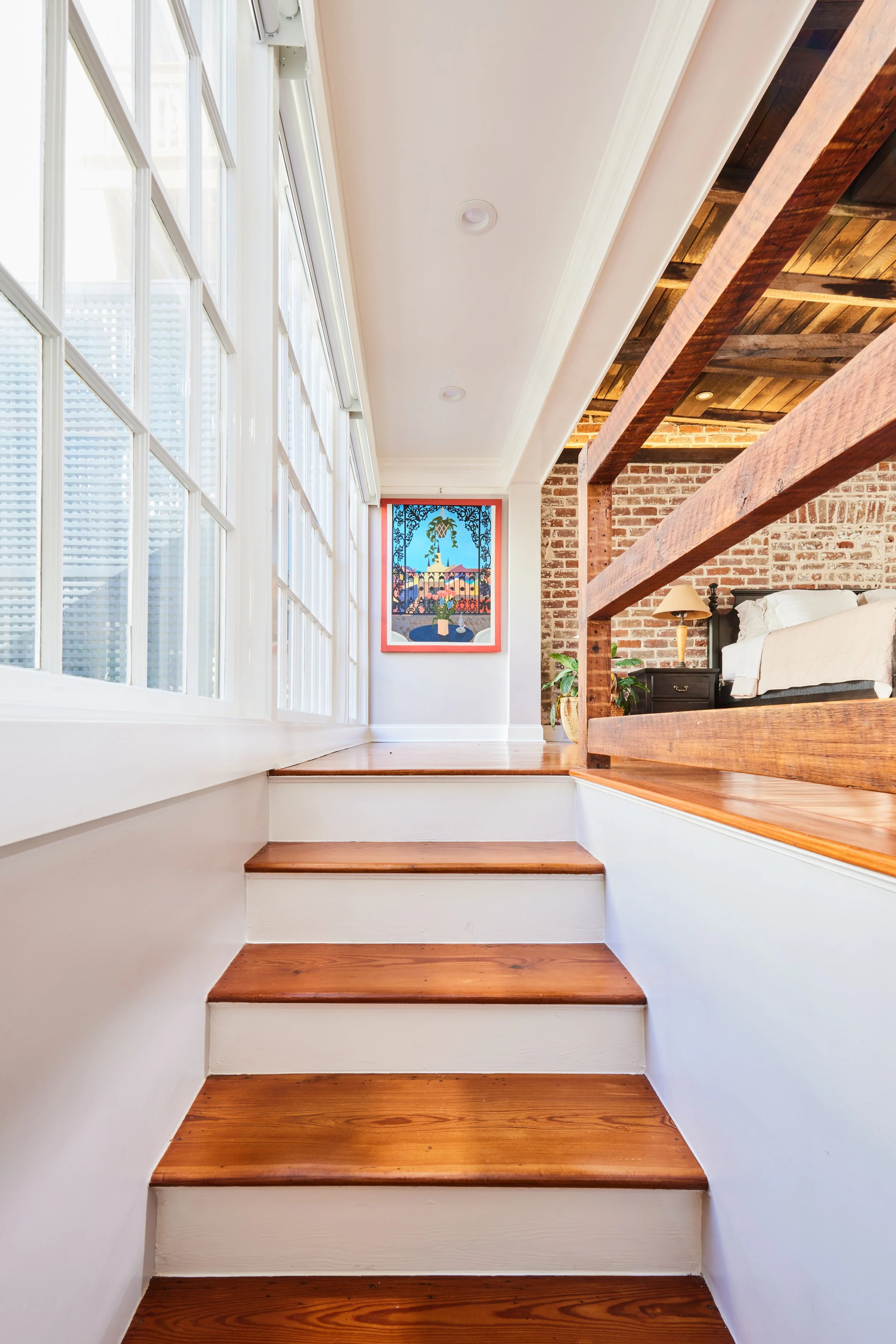 Wooden stairs leading up to a small landing area with white walls, a large window with multiple panes, a colorful picture on the wall, and part of a brick wall and bed visible in the background.