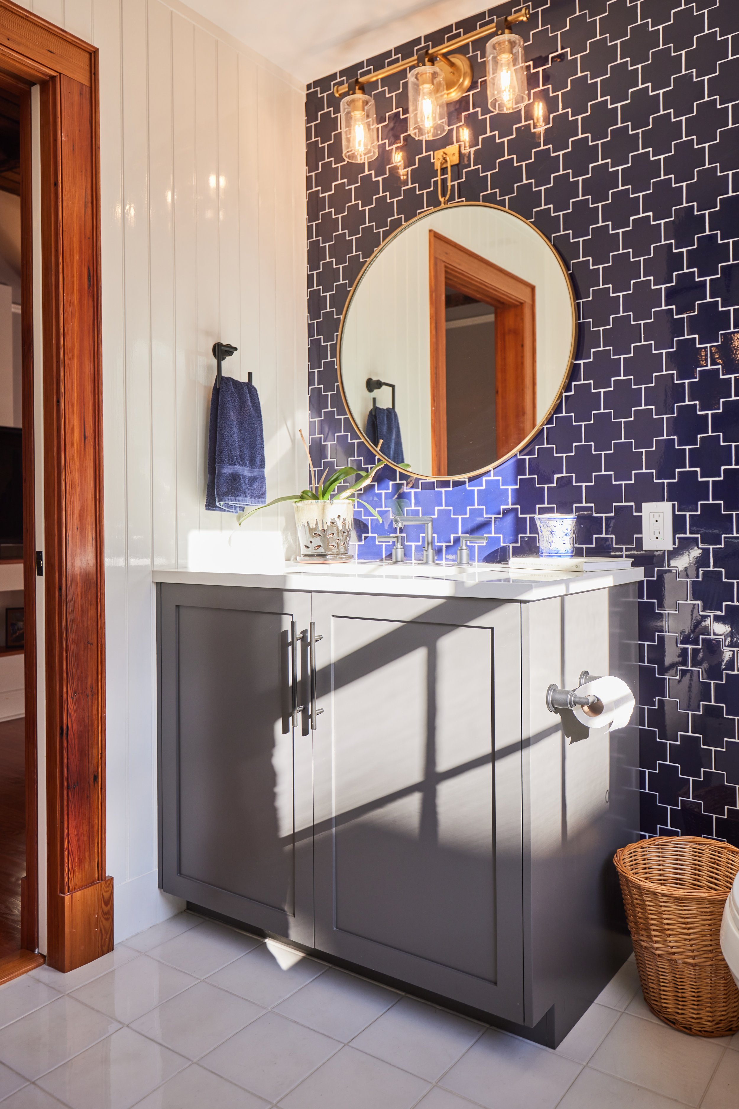 Bathroom vanity with a gray cabinet, white countertop, and a round mirror above. Blue towels hanging on wall hooks, potted plant, soap dispenser, decorative container, and a basket on tiled floor. Dark patterned wallpaper behind the mirror.
