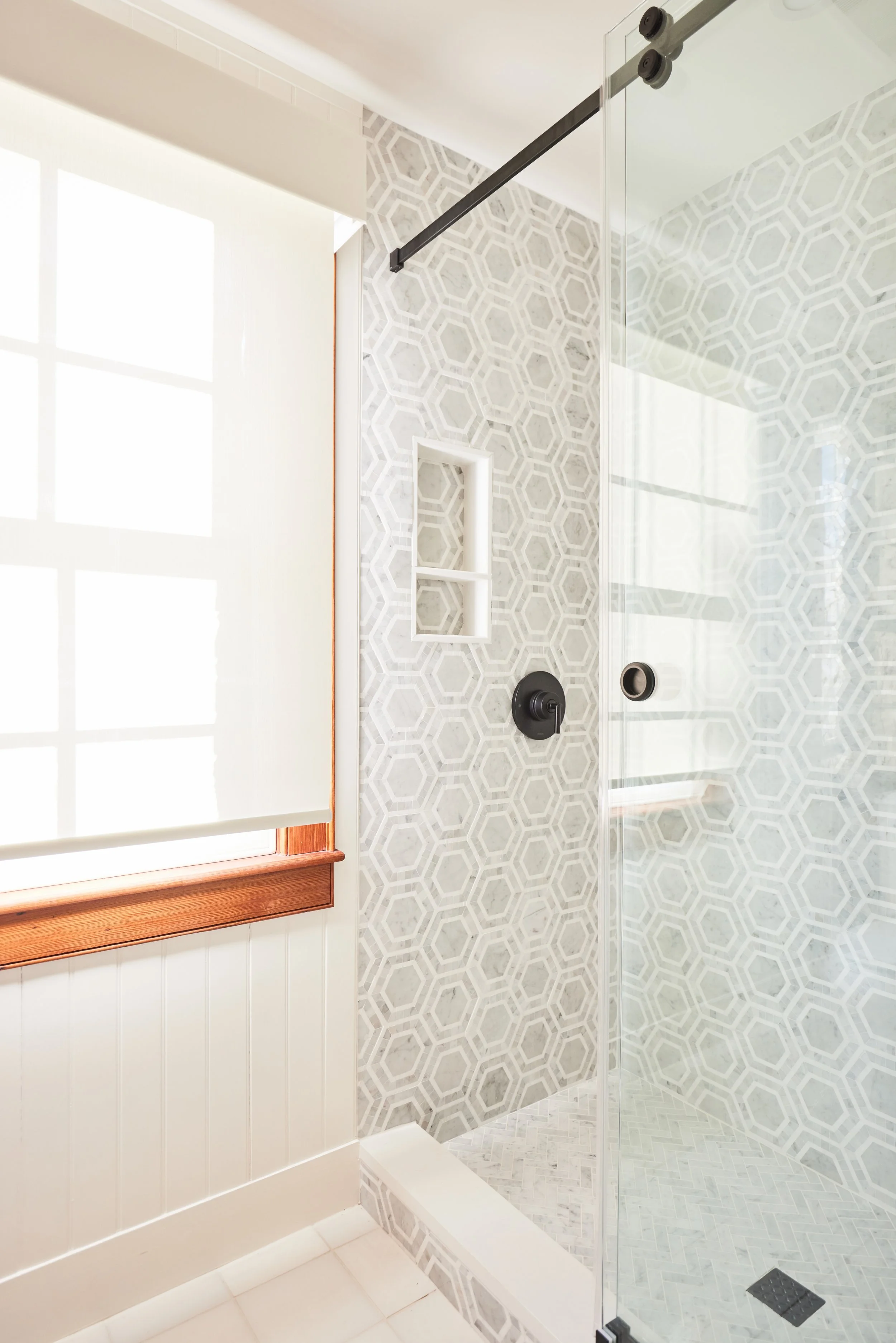 A modern walk-in shower with geometric patterned tiles, a black shower fixture, a built-in shelf, a glass door, a window with a white roller blind, and a wooden window frame.