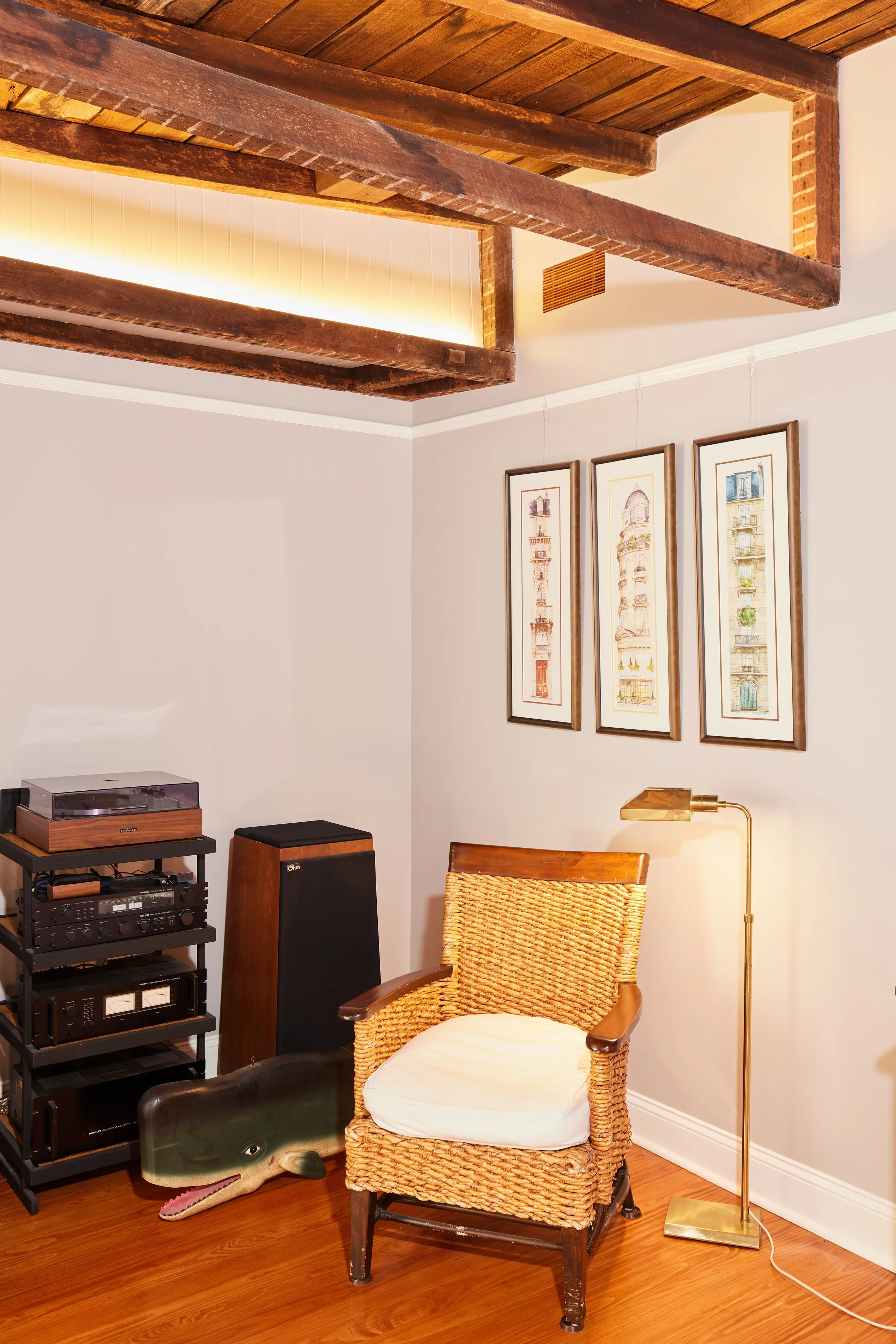 A cozy corner of a room with a wicker armchair with a white cushion, a tall gold floor lamp, three framed architectural sketches on the wall, wooden ceiling beams, a black audio equipment rack, a vintage turntable, a large speaker, and a decorative dolphin figurine on the floor.
