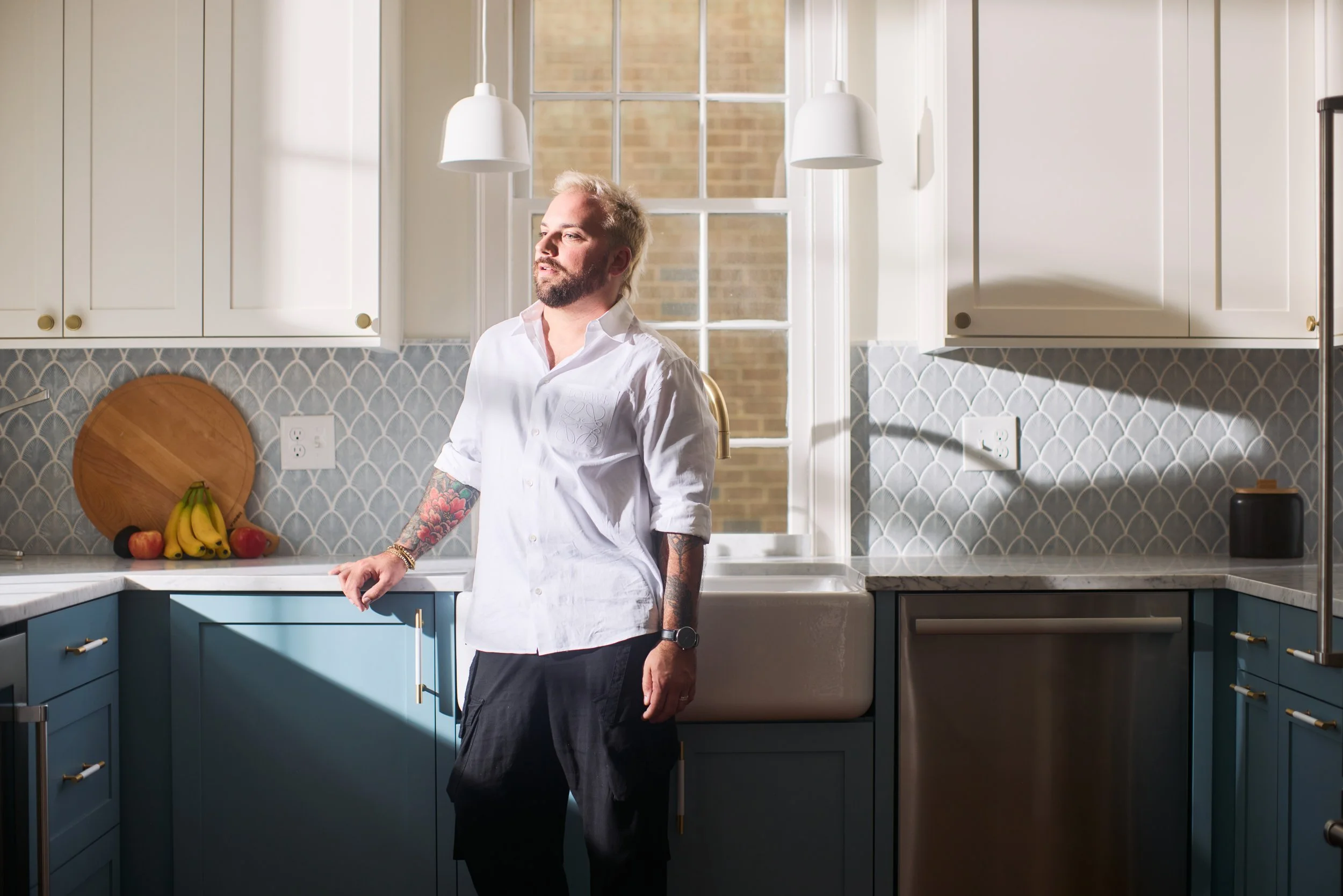 A man with tattoos standing in a bright kitchen near a window with sunlight, wearing a white shirt and black pants.
