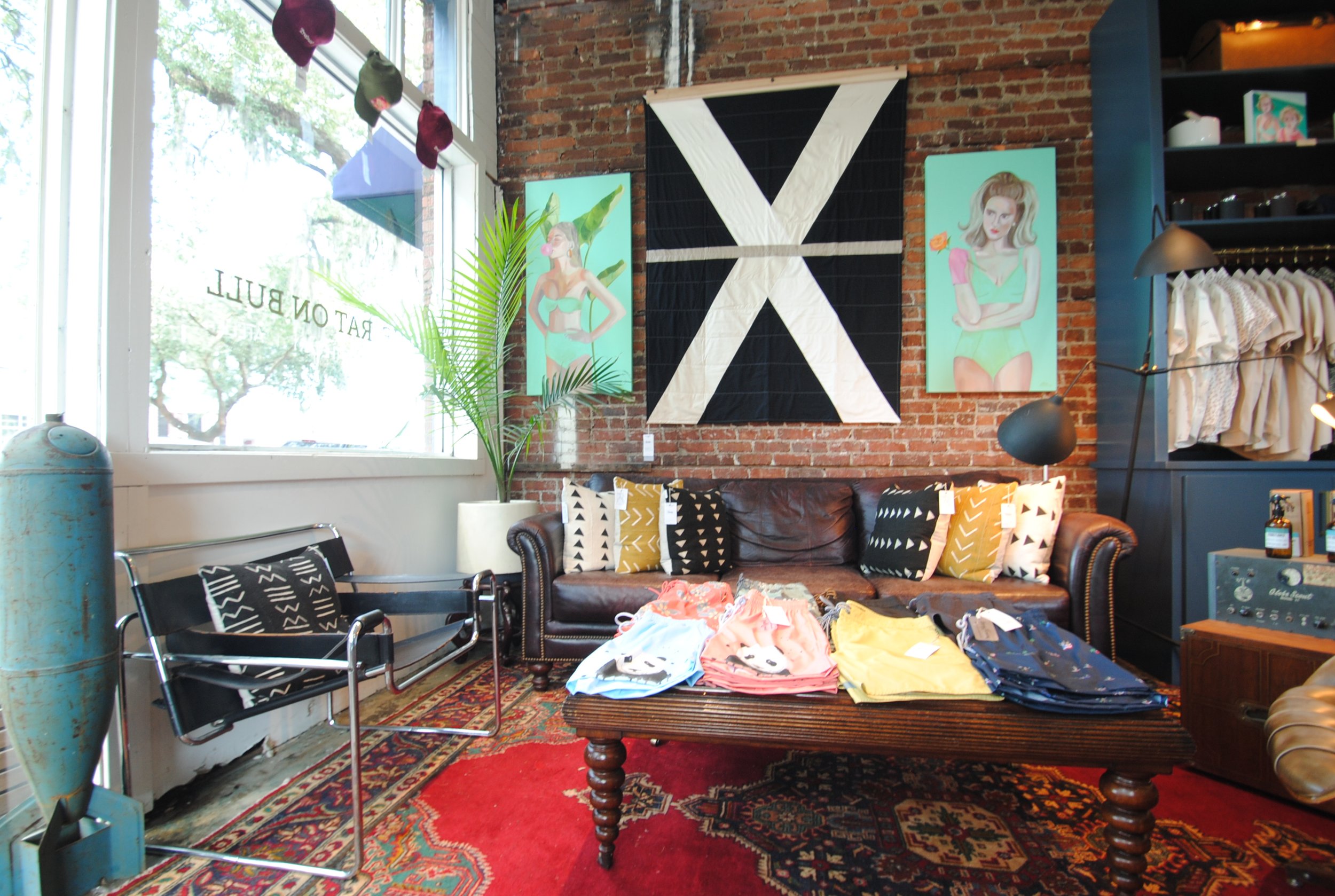 Interior of a vintage-style store with a leather sofa, a wooden coffee table displaying folded clothes, colorful patterned pillows, a modern black and chrome chair, a large potted plant, artwork on brick walls, and a display of clothing.