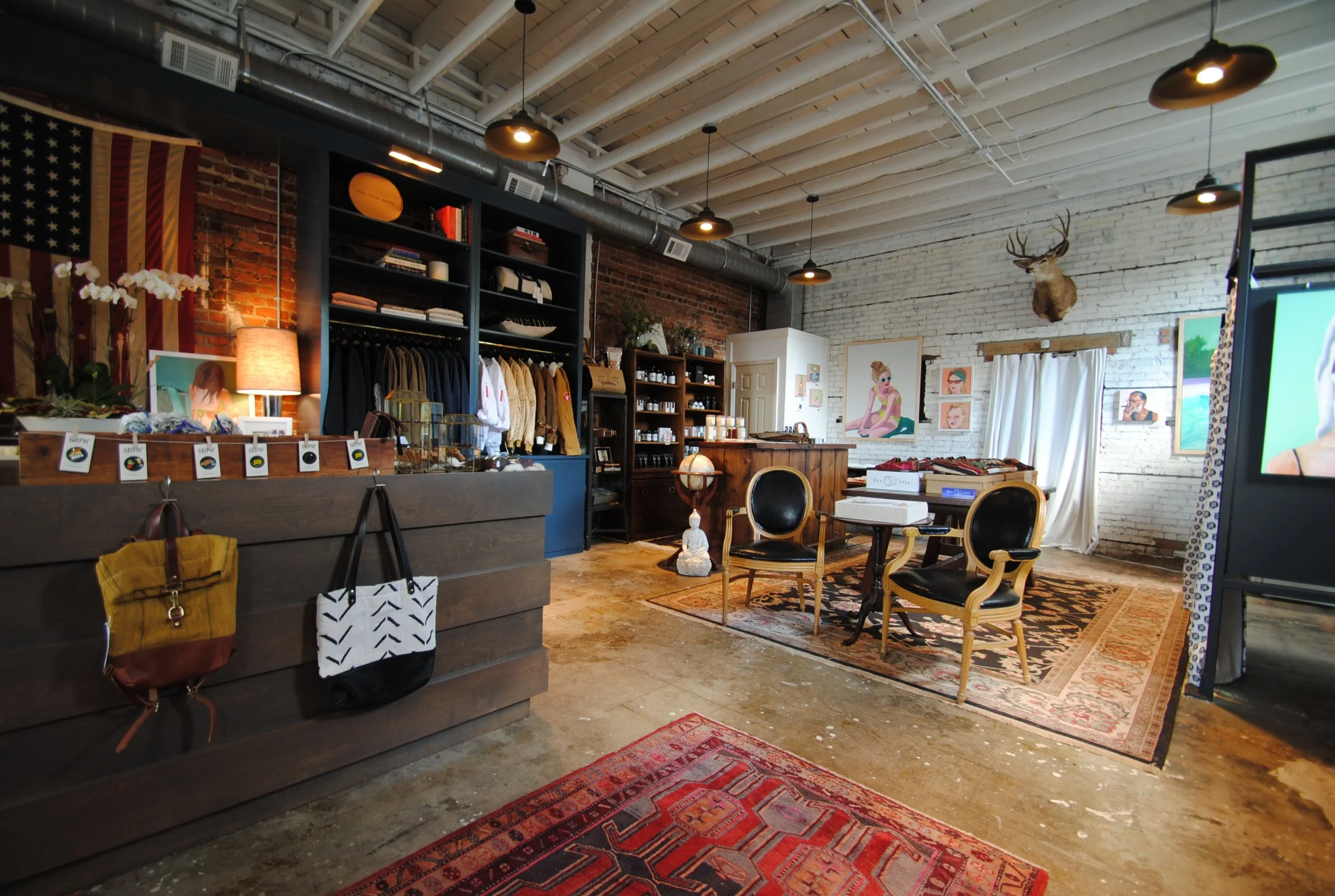 Interior of a boutique store with clothing on display, artwork on the walls, a deer head mounted on the white brick wall, and a seating area with chairs and a rug.