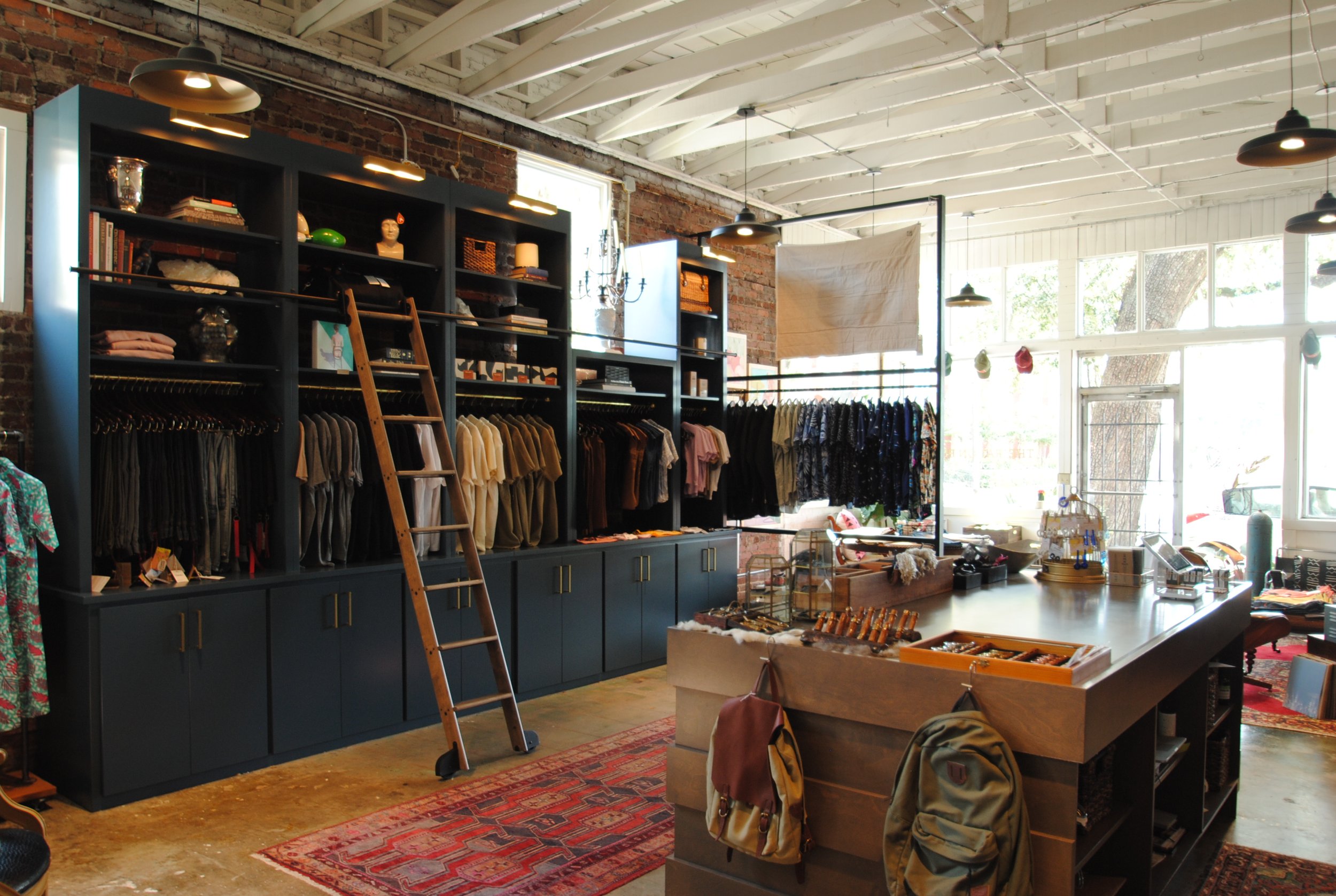 Interior of a boutique clothing store with dark shelving units filled with clothing, accessories, and decorative items, a wooden ladder leaning against the shelf, a central checkout counter with items on display, and large windows allowing natural light to illuminate the space.