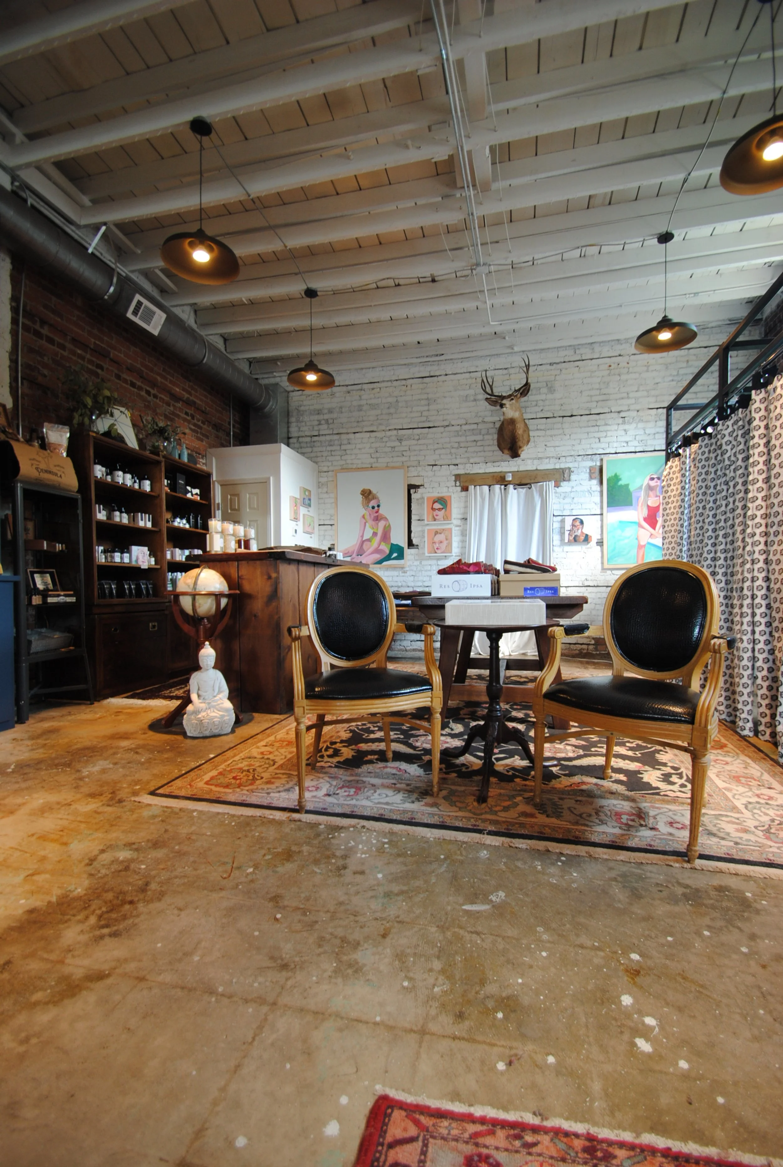 Interior of a cozy room with vintage chairs, paintings on a white brick wall, a mounted deer head, and eclectic decor. The space has a rustic wood ceiling, exposed ductwork, and a patterned rug on the concrete floor.