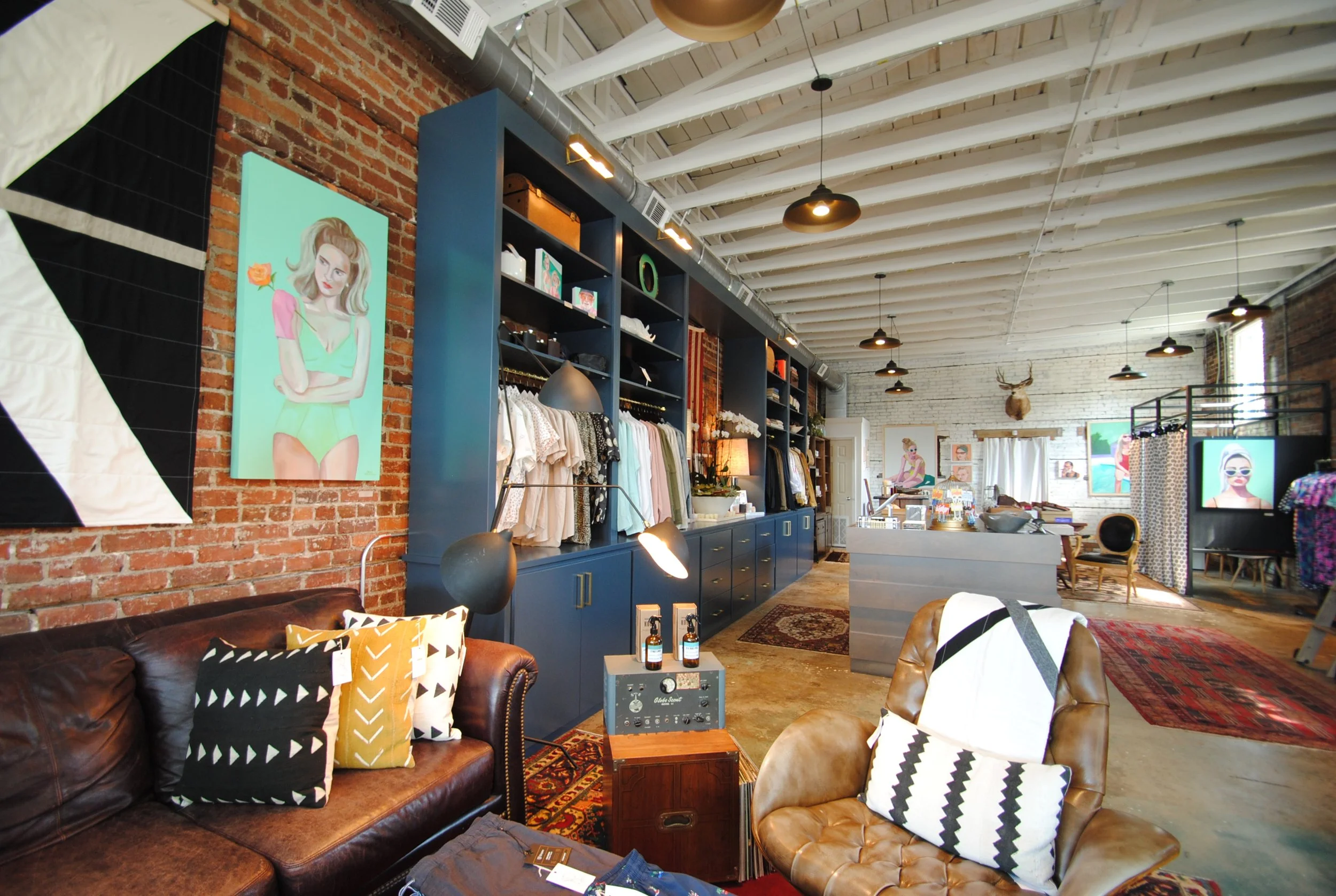 Inside a boutique store with exposed brick walls, wooden floors, and modern decor. There are artwork on the walls, clothing on blue shelving, a sofa with patterned pillows, and a leather chair. The ceiling has white beams and hanging lights.
