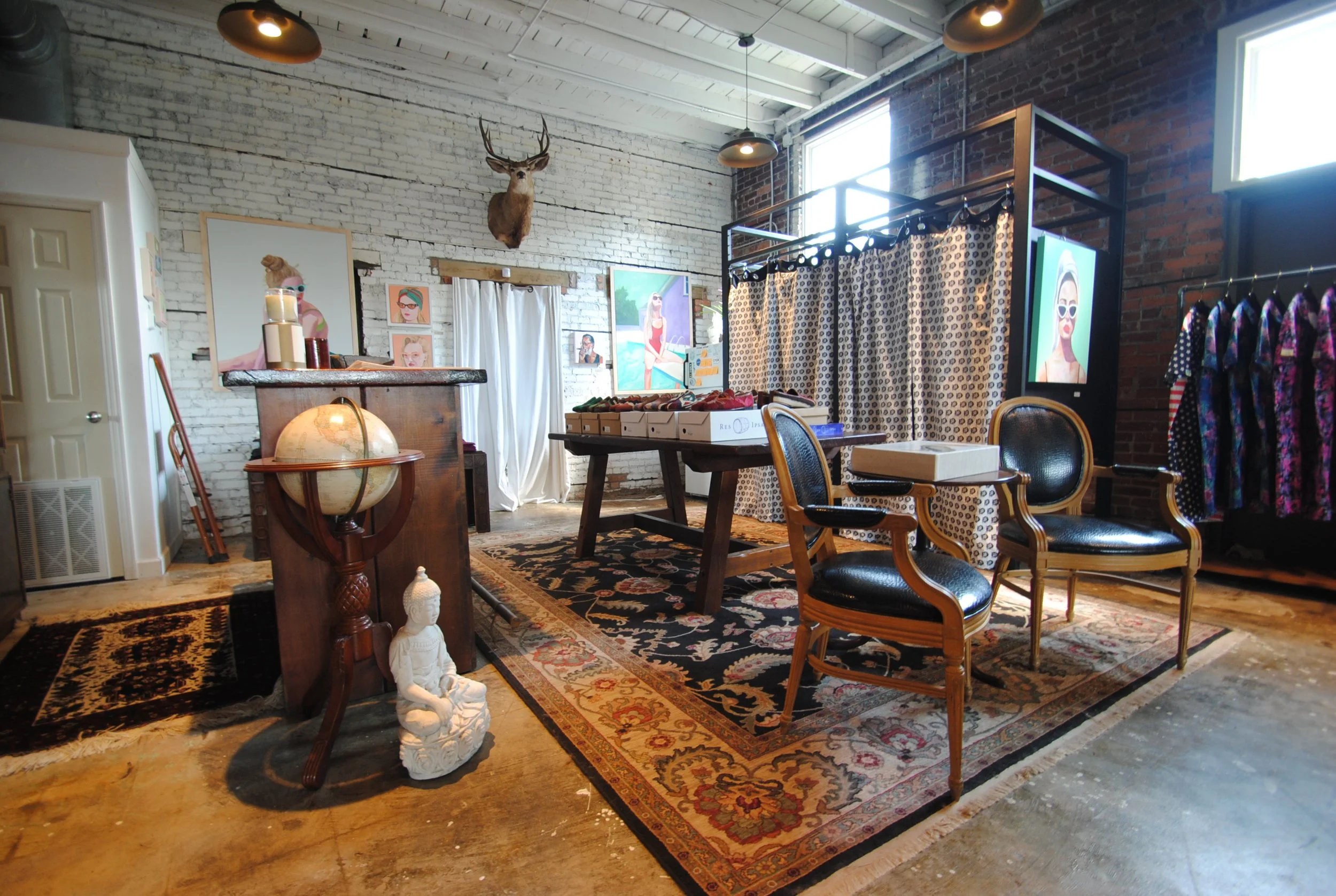 Interior of a boutique or art studio with vintage furniture, artwork, decorative items, clothing rack, and a mounted deer head on a white brick wall.