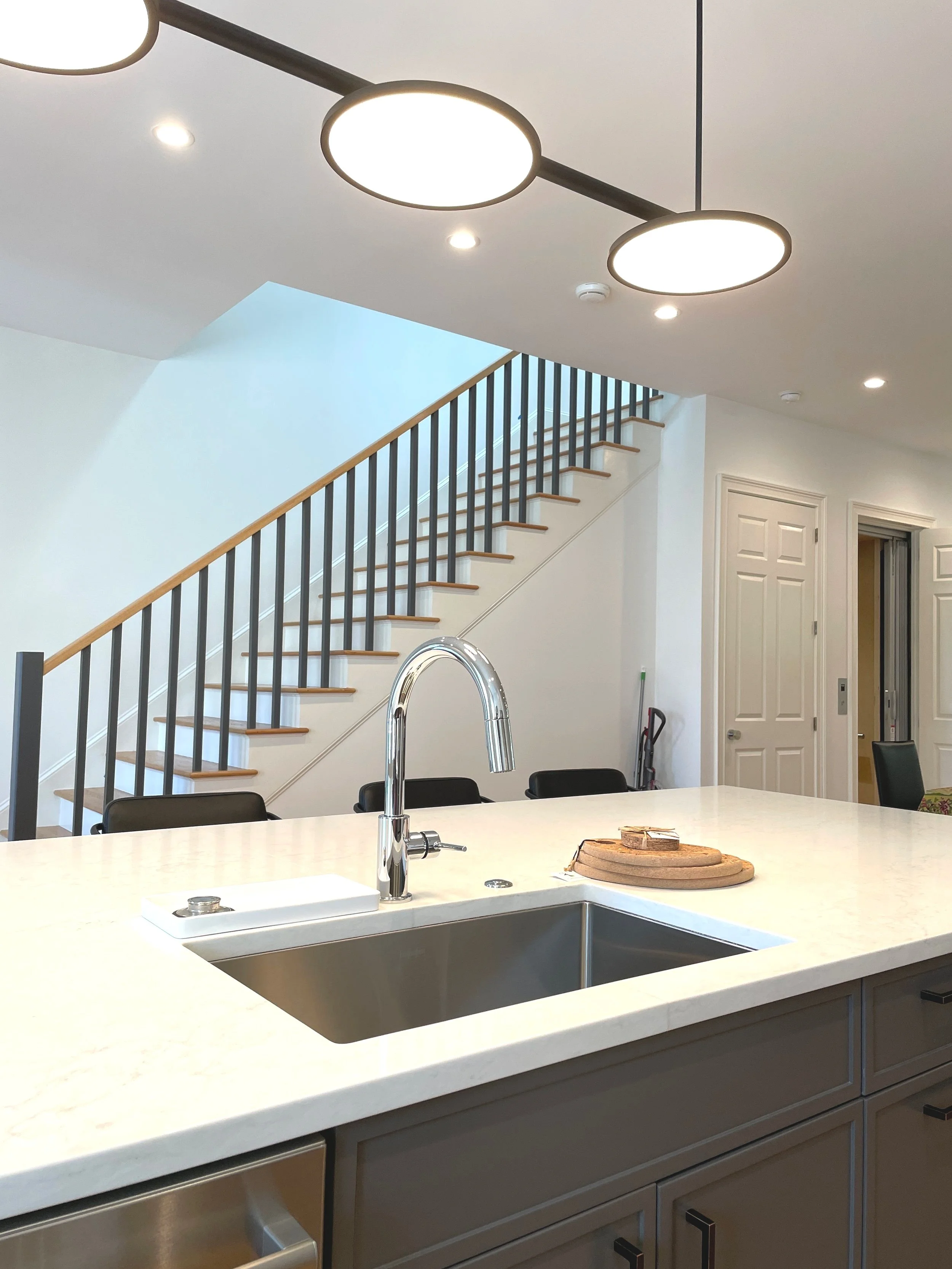 Modern kitchen with a white island counter and stainless steel sink, a chrome faucet, and black chairs. In the background, a staircase with wooden steps and a black railing leads upstairs. The ceiling has circular light fixtures.