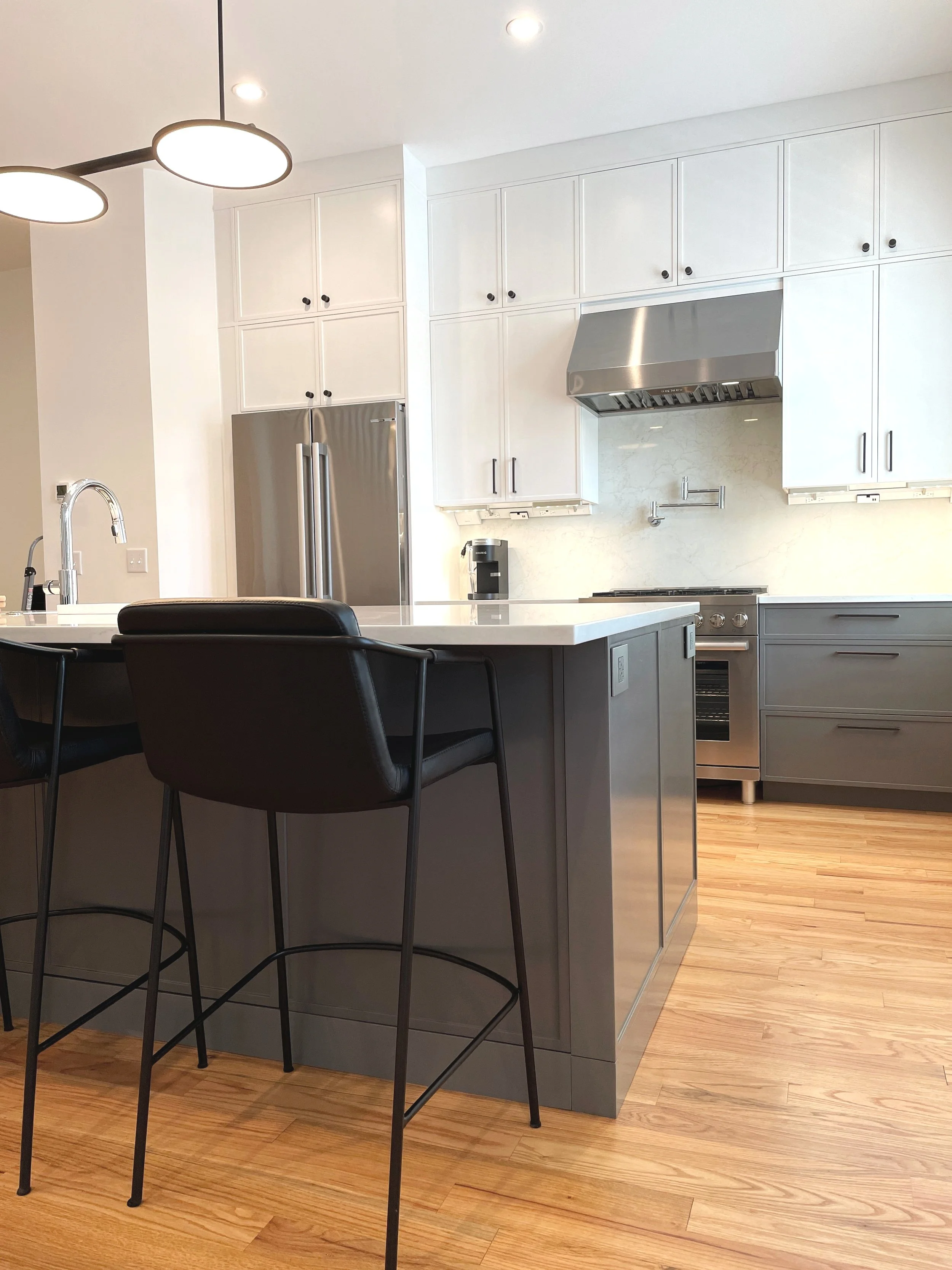 Modern kitchen with white cabinets, stainless steel appliances, gray island with black chairs, and wooden flooring.