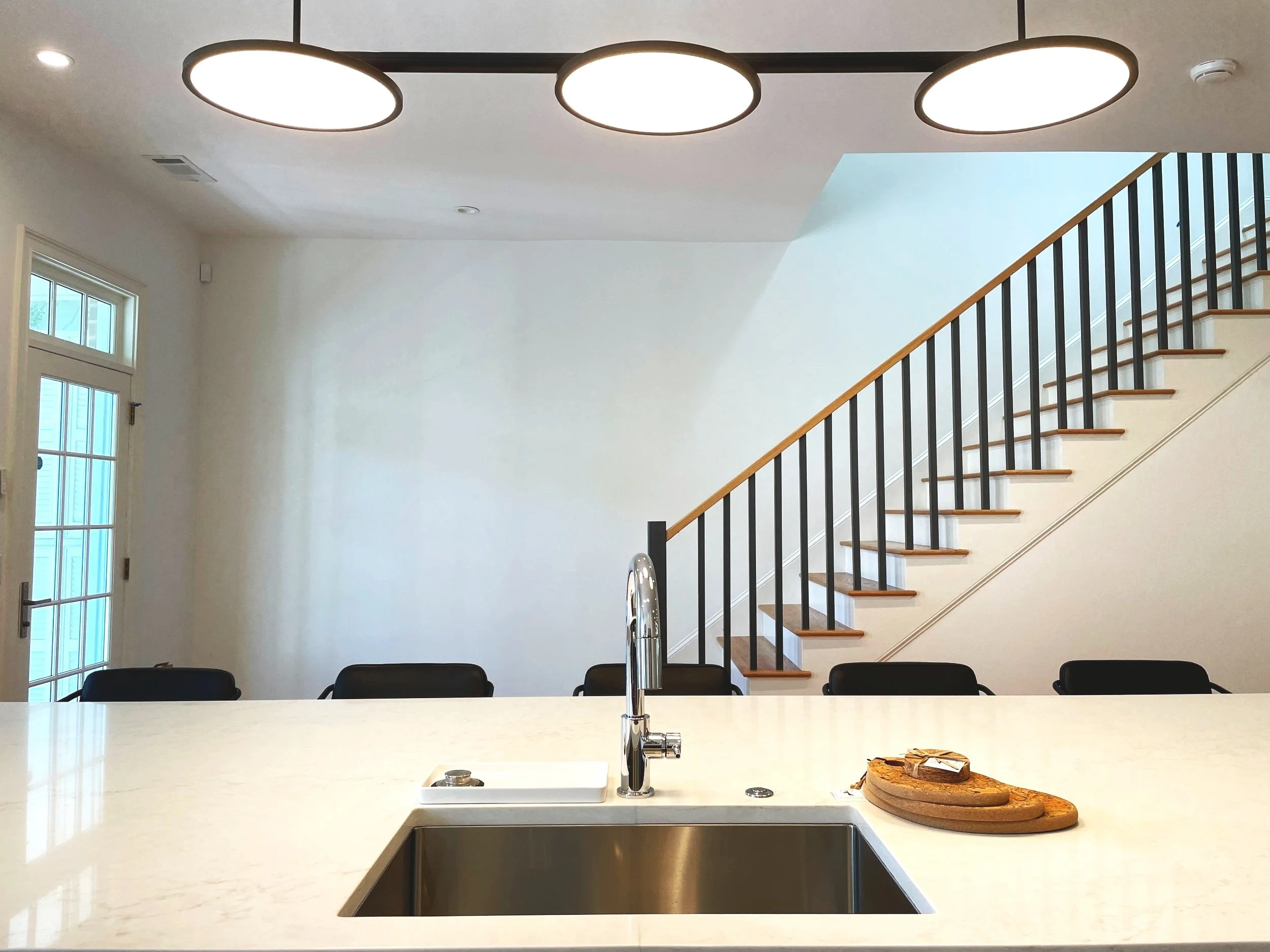 Modern kitchen with a white countertop, black chairs, staircase with wooden steps and black railing, large circular ceiling lights, side door with glass panes.
