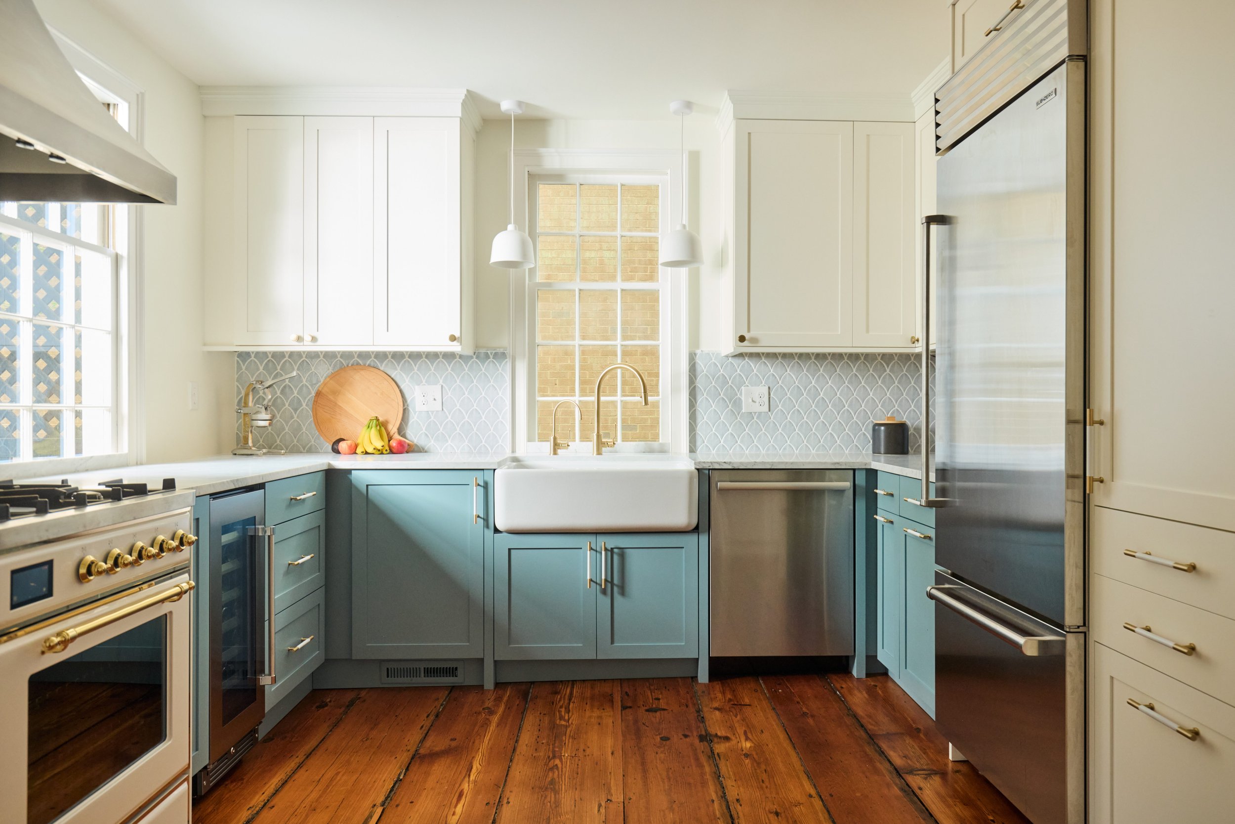 Bright kitchen with white upper cabinets, blue lower cabinets, a farmhouse sink under a window, stainless steel appliances, and hardwood floors.