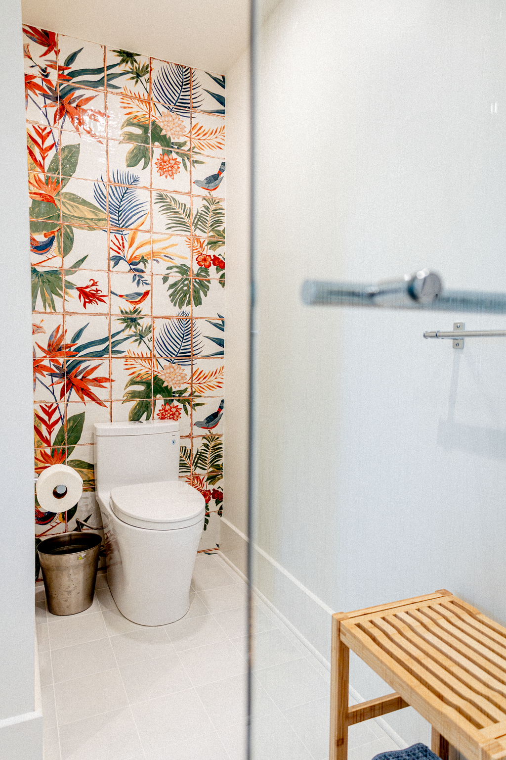 Bathroom with tropical patterned tiles behind the toilet, a showerhead on the right, a wooden bench, and a metal waste bin.