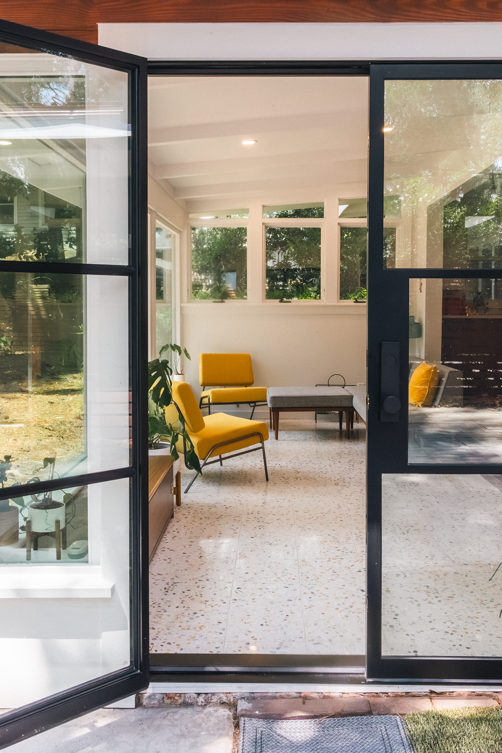 Interior view of a sunroom with yellow chairs, a small table, potted plants, and large windows, seen through an open glass door.