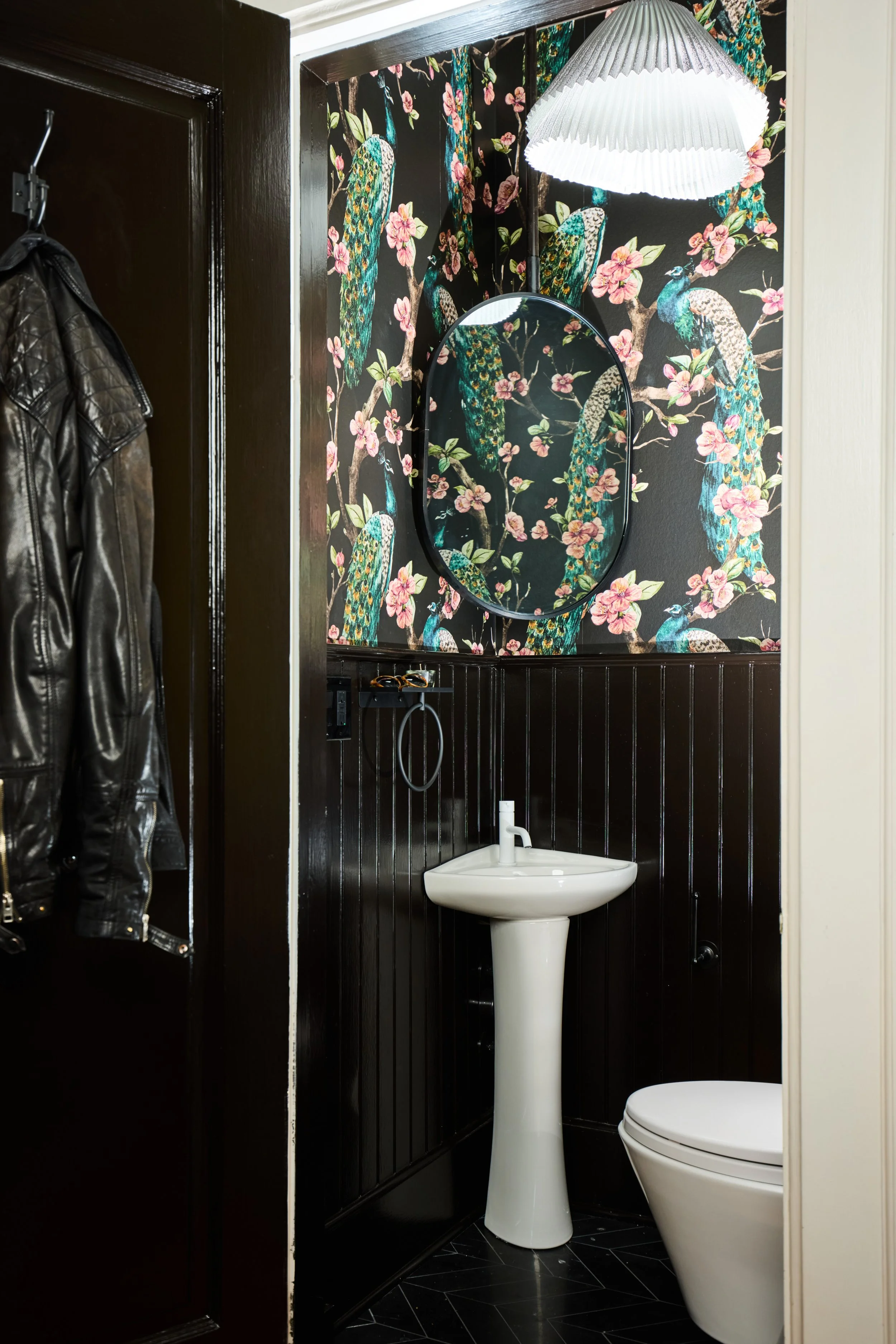 A small bathroom with floral peacock wallpaper, a white pedestal sink, a mirror, and a hanging ceiling light. Part of a black leather jacket is visible on the left side.