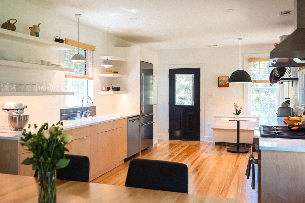 Bright, modern kitchen with wooden floors, white walls, open shelving, stainless steel appliances, a black door, and large windows letting in natural light.