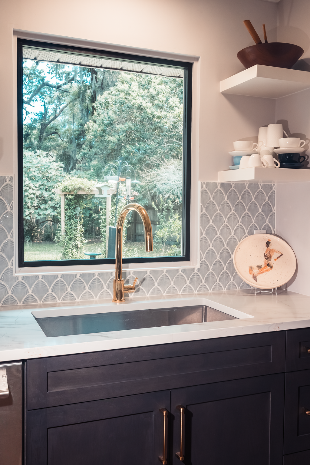 Kitchen with a window view of a lush green backyard, black cabinets with gold handles, a white marble countertop, a gold faucet, and shelves holding cups, bowls, and a decorative plate with a gymnast illustration.