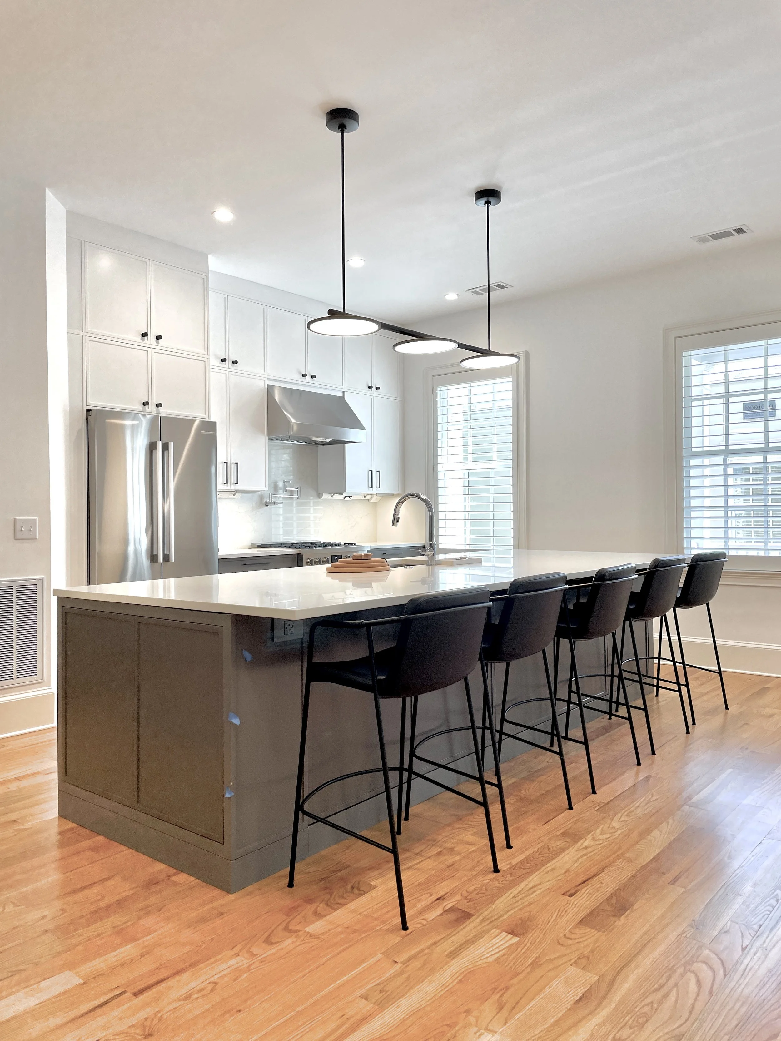 Modern kitchen with white cabinets, stainless steel refrigerator, wooden floor, pendant lights above a kitchen island with black chairs, and large windows with blinds.