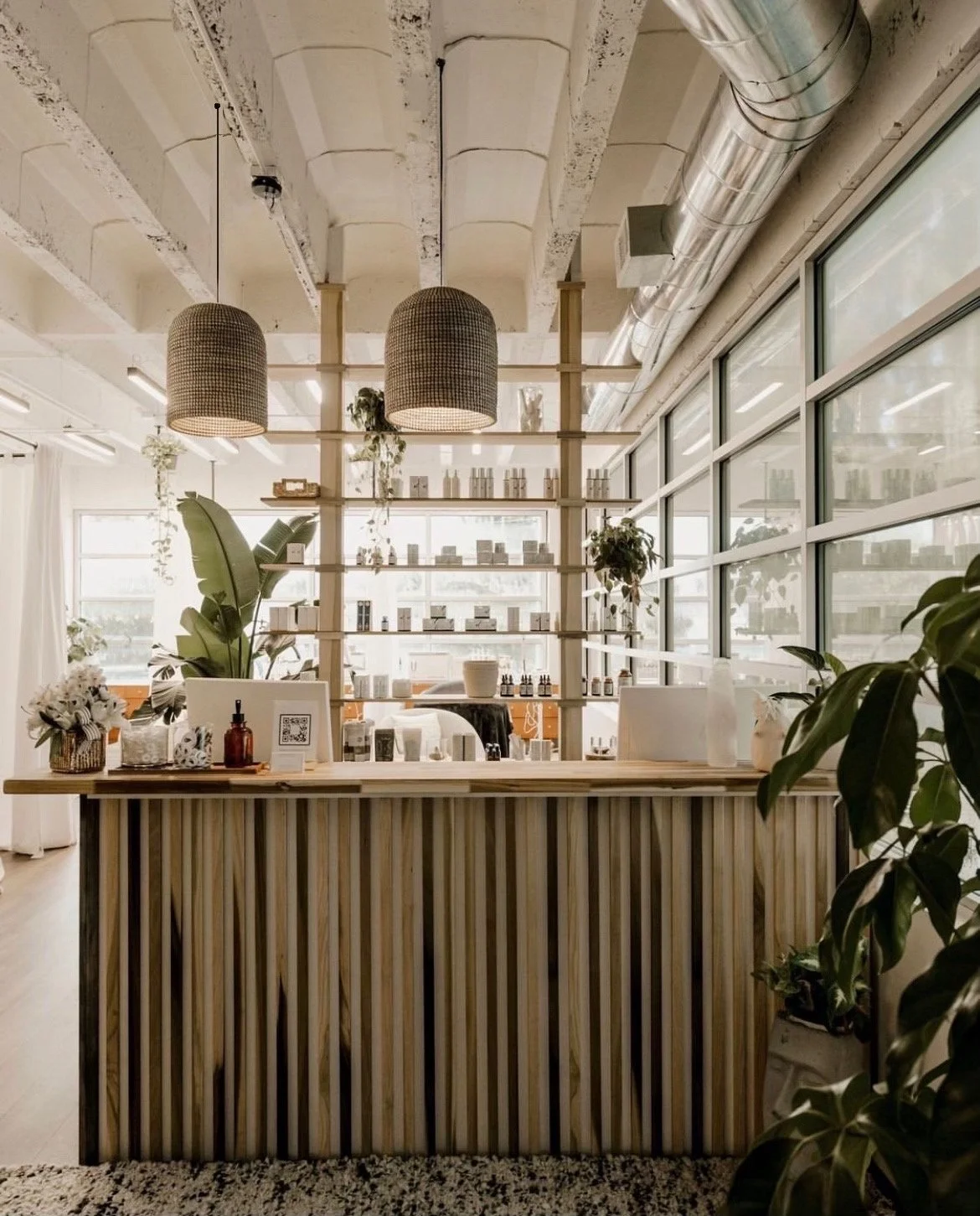 Interior of a modern boutique or skincare store with wooden reception desk, shelves with skincare products, large windows, hanging woven lamps, and various plants.
