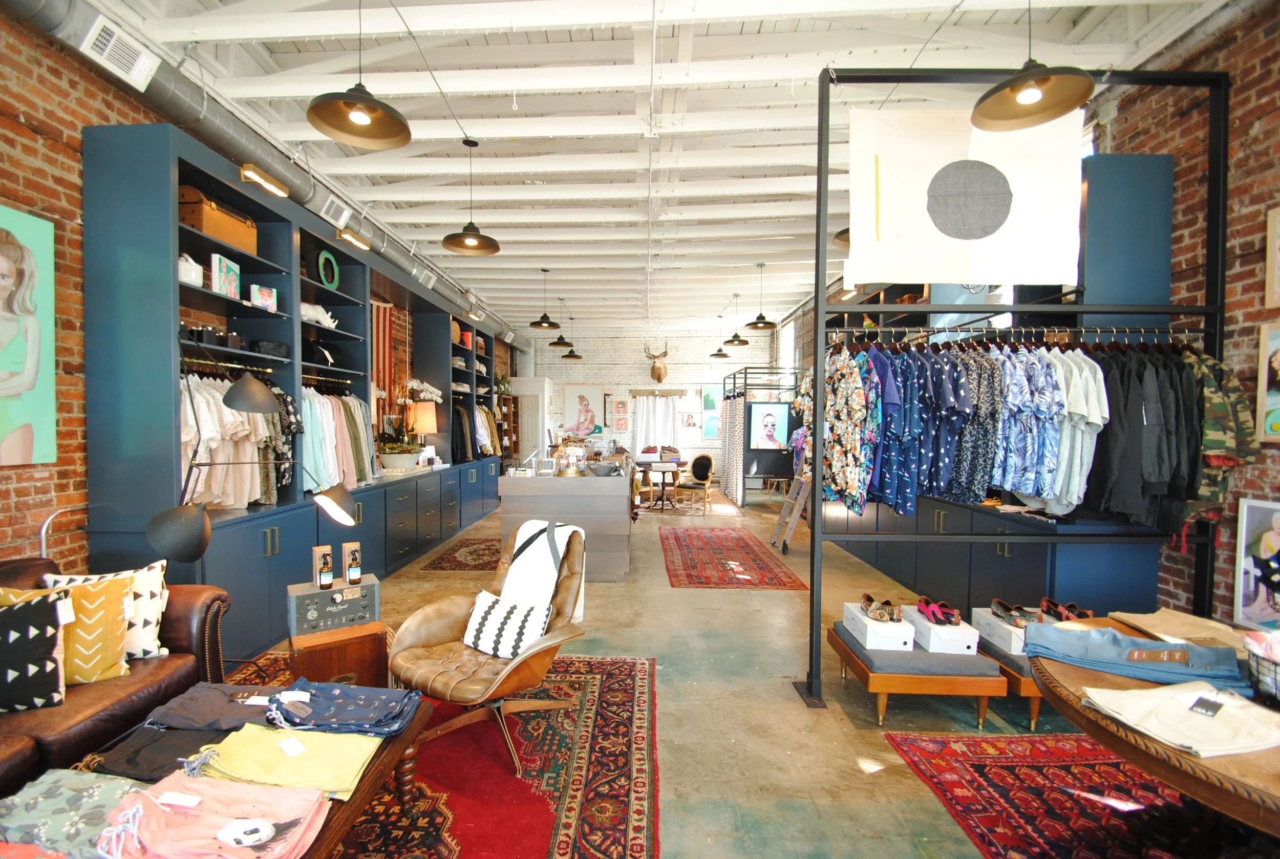 Interior of a stylish clothing boutique with exposed brick walls, wooden floors, and colorful paisley and traditional rugs. Racks of men's and women's apparel, shelves with accessories, and a cozy seating area with pillows and lamps are visible.