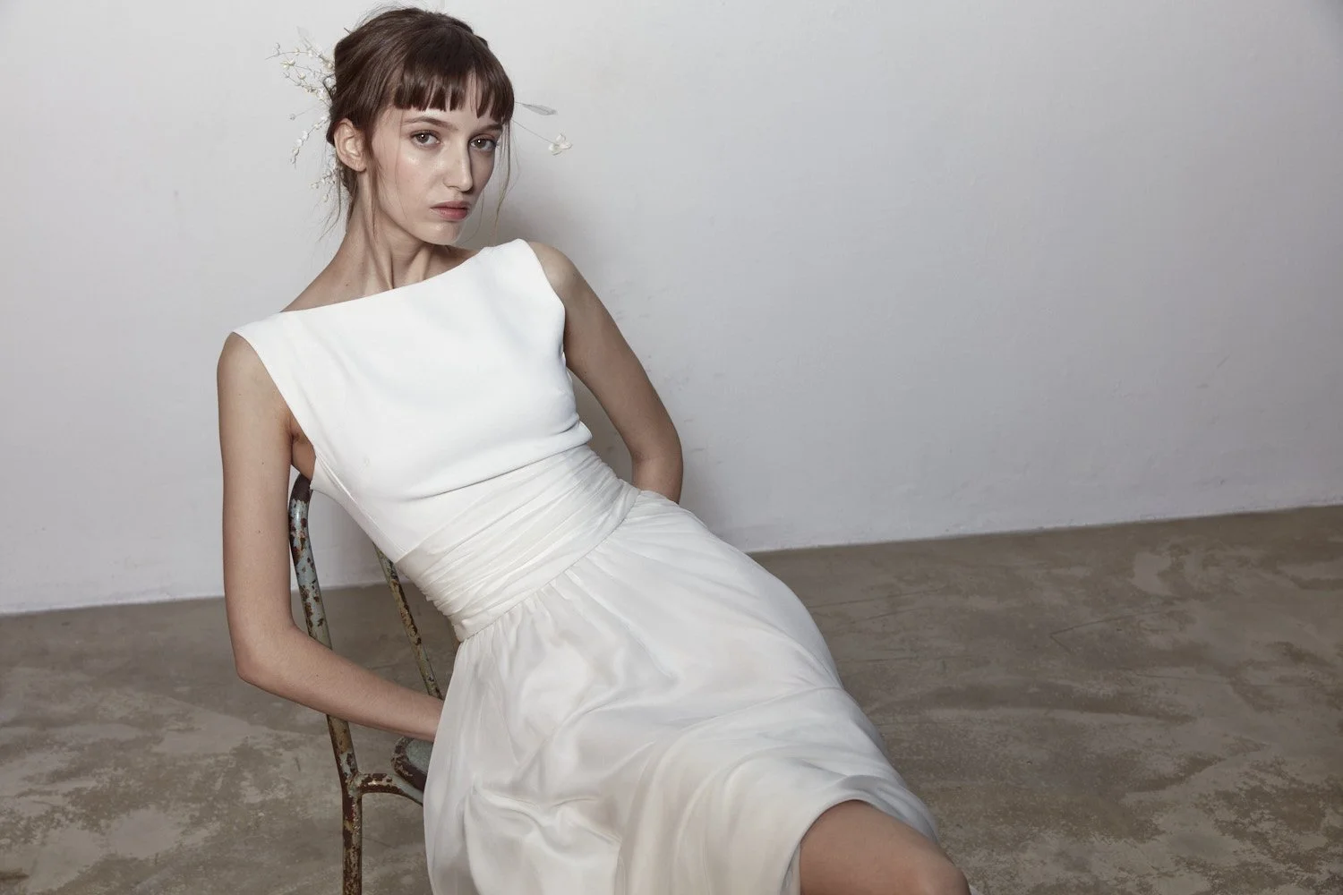 Una donna con capelli castani, abbigliamento bianco, seduta su una sedia in un ambiente minimalista con pareti bianche e pavimento in cemento. Porta un vestito bianco elegante e un accessorio floreale tra i capelli.