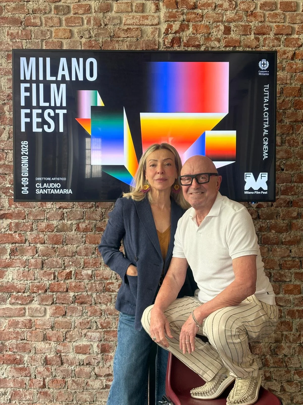 Due persone, una donna e un uomo, si trovano davanti a un muro di mattoni rossi, in piedi e seduto. Sullo sfondo, un grande schermo con il logo del Milano Film Fest e dettagli della manifestazione. La donna indossa una giacca blu e pantaloni blu, l'uomo indossa una maglietta bianca, pantaloni a righe e occhiali neri.