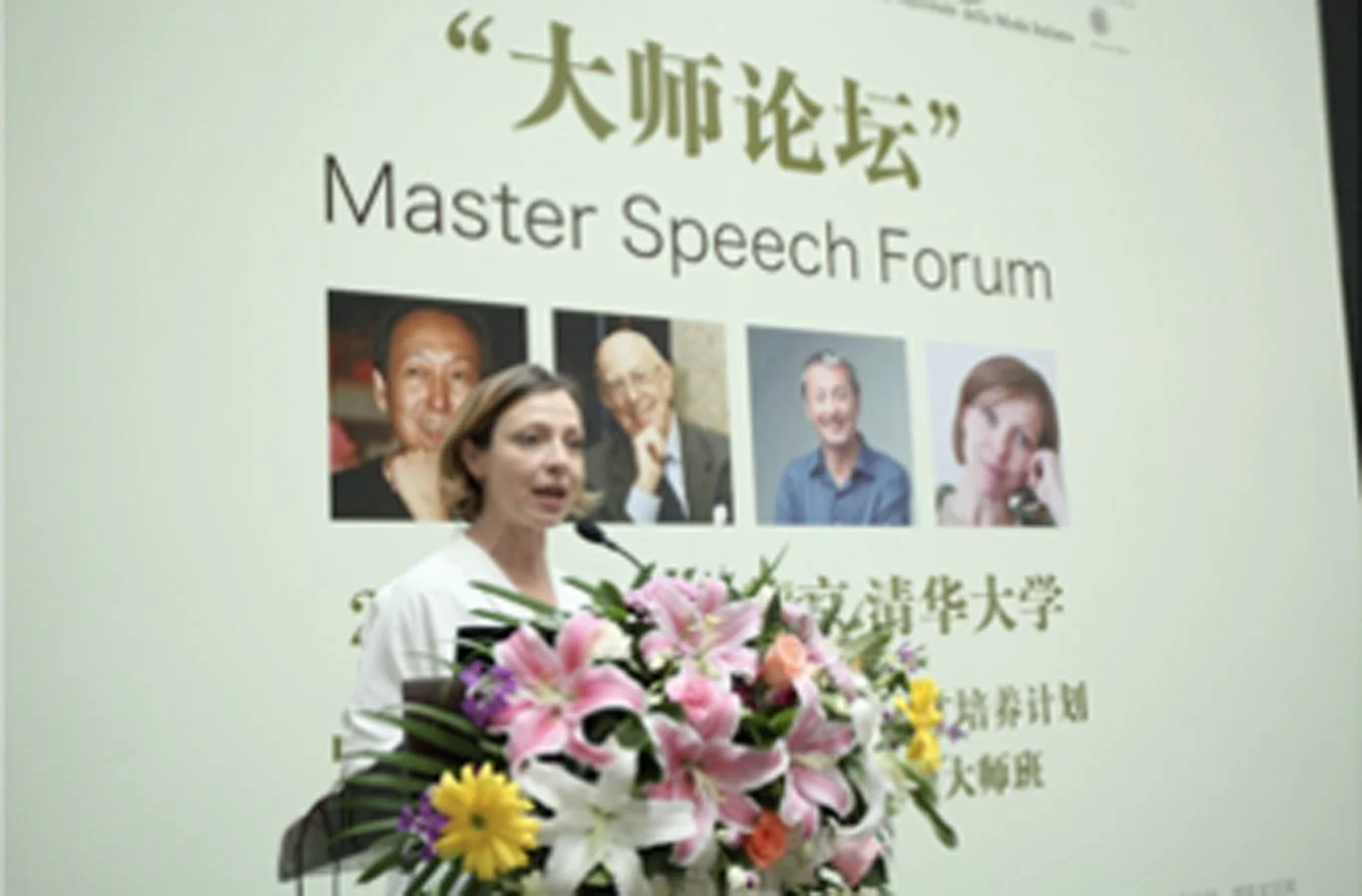 Donna che parla a un microfono durante il Forum di Oratoria, con uno schermo dietro che mostra immagini di altri relatori e testo in italiano e inglese, decorato con un mazzo di fiori.