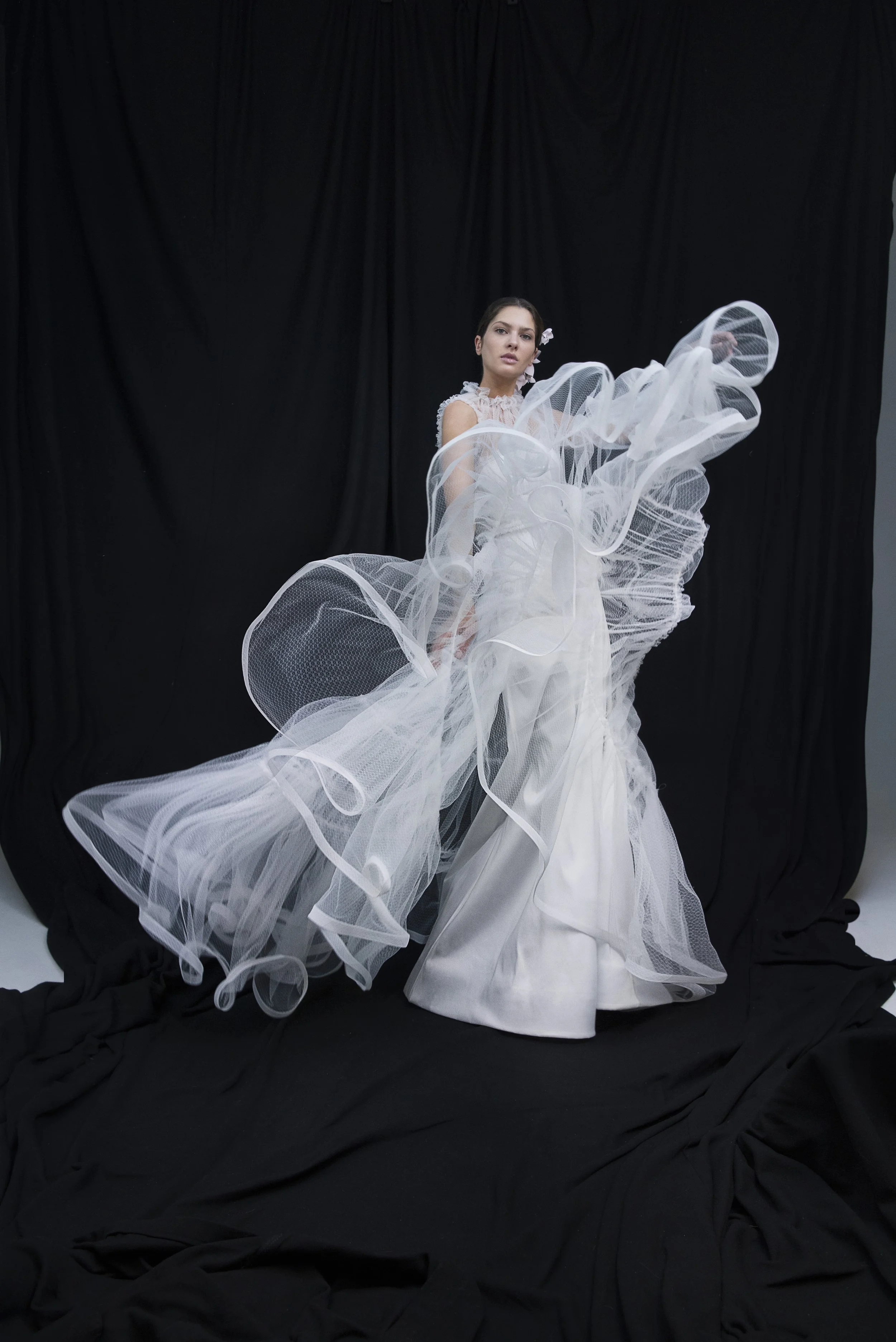 Una donna indossa un abito da sposa bianco con dettagli di tessuto a rete e volant, posizionata davanti a uno sfondo nero.