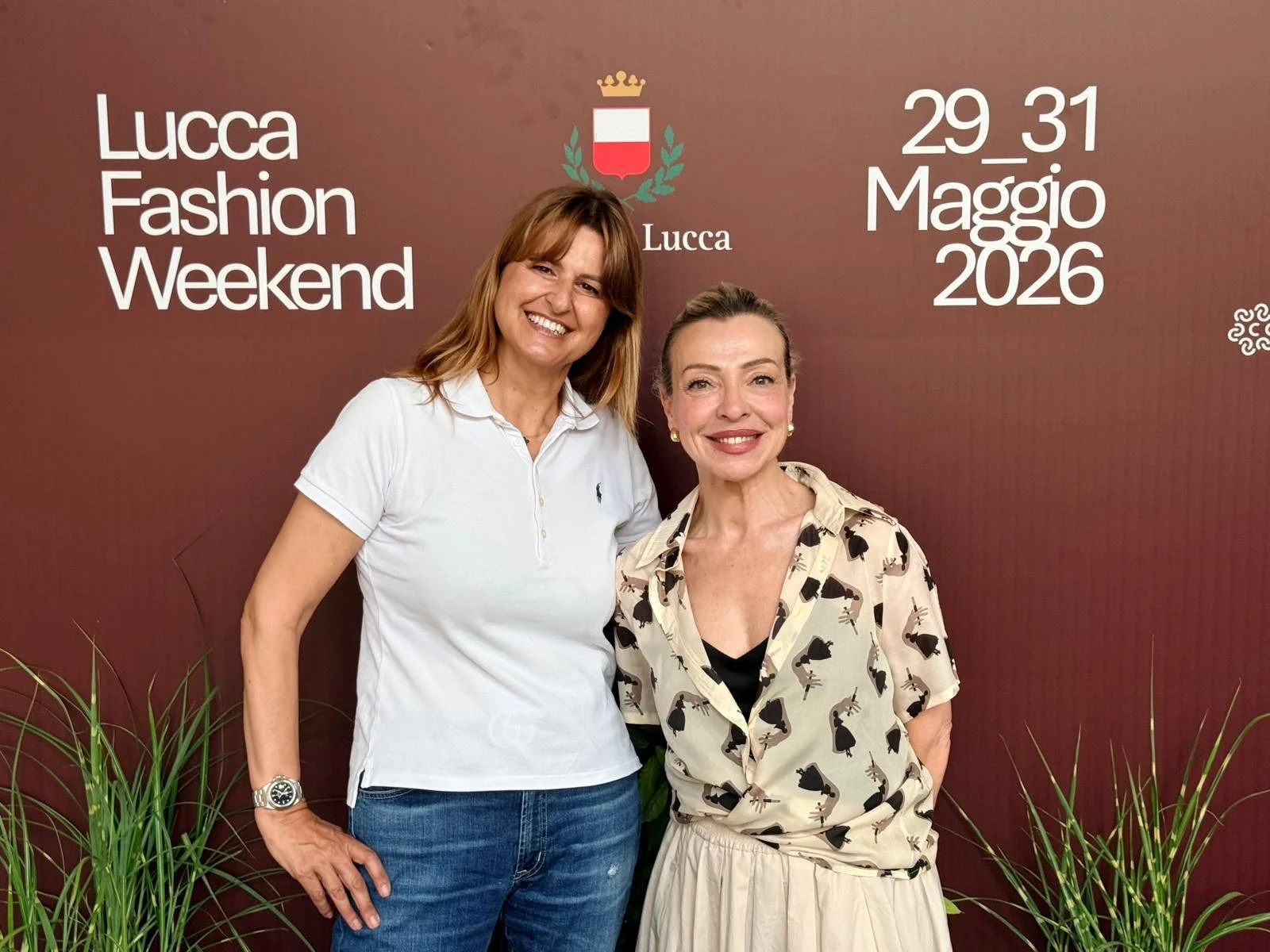 Due donne sorridenti davanti a uno sfondo con testo e simboli, in occasione del Lucca Fashion Weekend, tenutosi dal 29 al 31 maggio 2026.