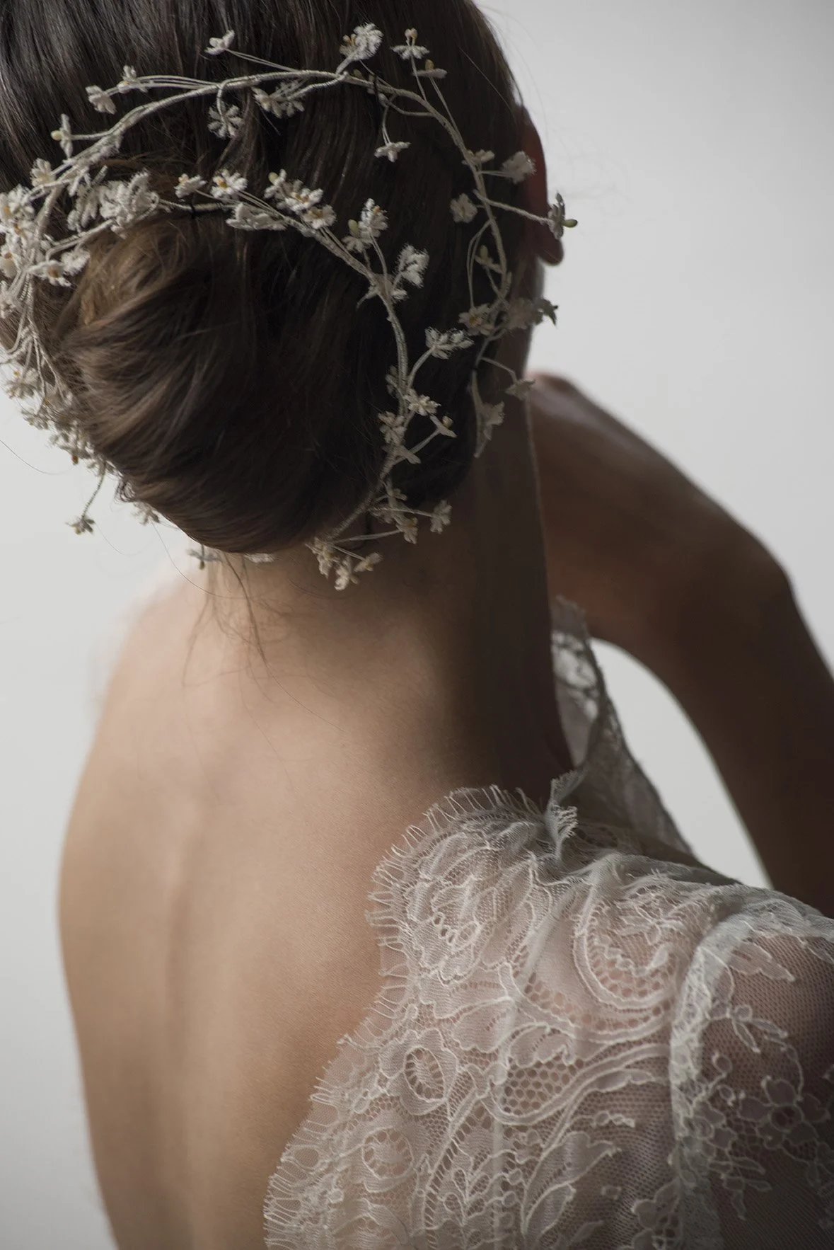 Dettaglio di una donna con capelli acconciati in uno chignon, decorata con una corona di fiori secchi, indossa un abito di pizzo morbido e semi trasparente, in una posa dallo stile delicato e romantico.