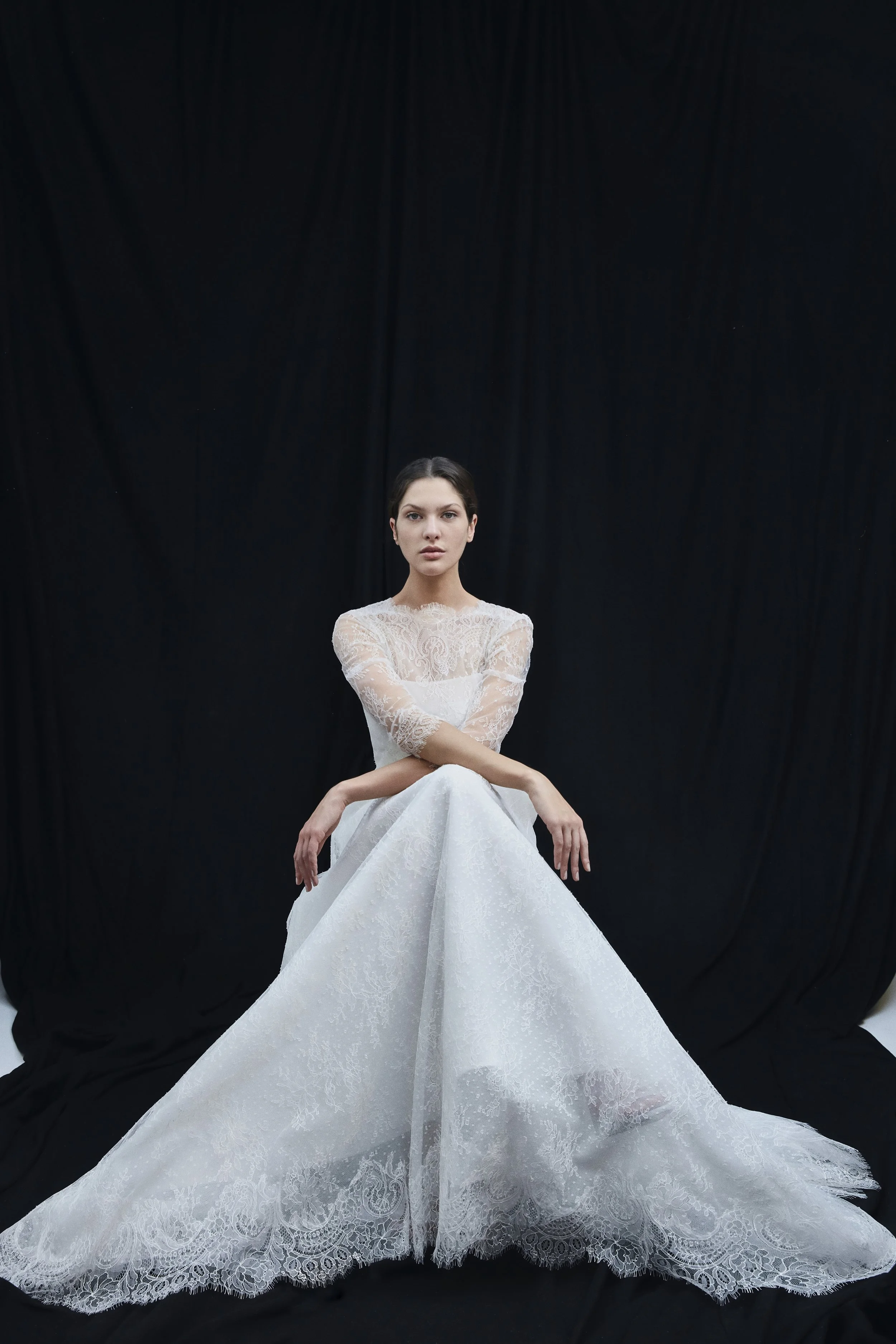 Donna con abito da sposa bianco in pizzo, seduta con le braccia incrociate davanti a uno sfondo nero.