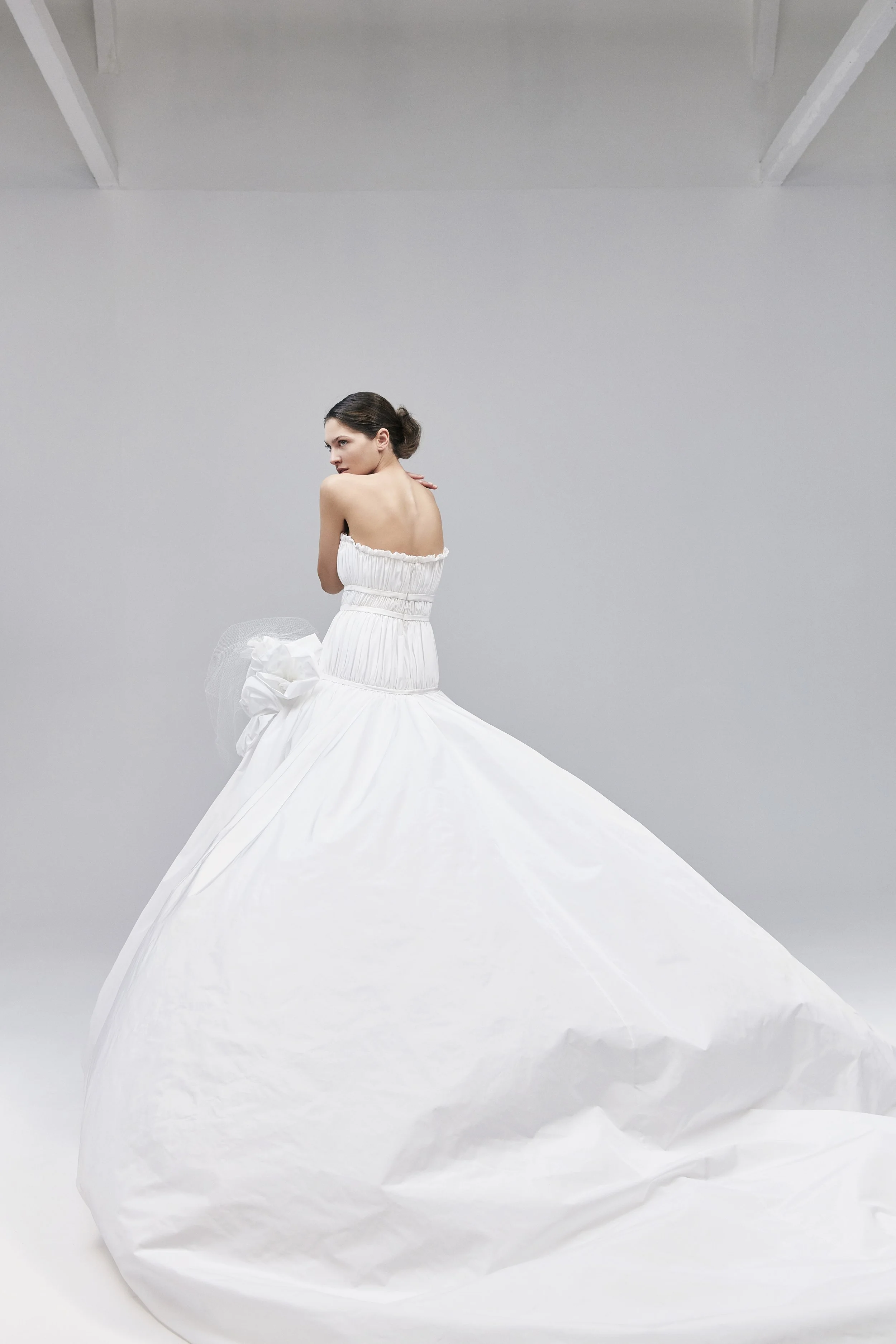 Una donna vestita con un abito bianco lungo e svolazzante si trova in uno spazio bianco e minimale, con le mani appoggiate alle spalle e guarda di lato.