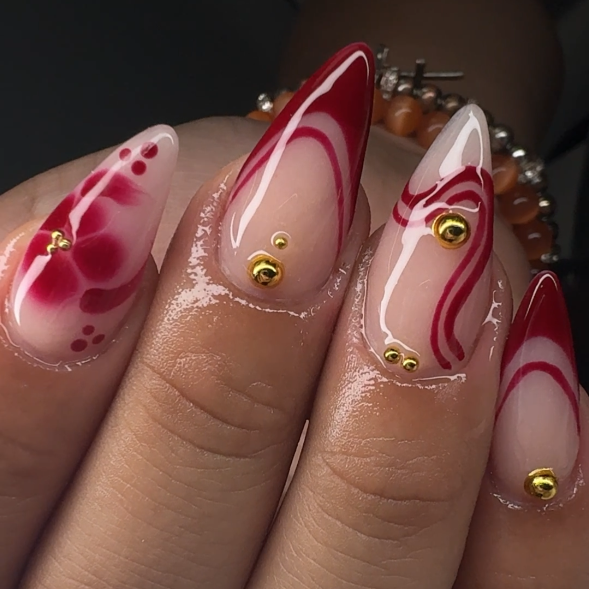 Close-up of a hand with elaborately painted, long, almond-shaped nails featuring red and white swirl and dot designs, decorated with small gold beads.