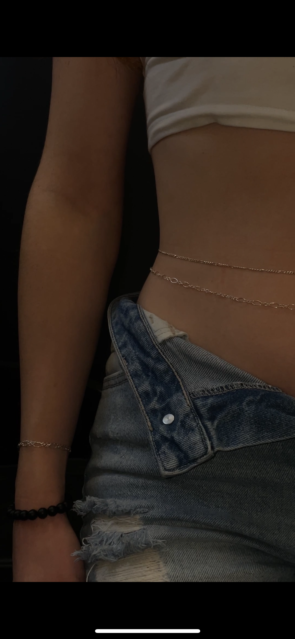 Close-up of a person lying down, showing their torso and partially unbuttoned denim shorts, a white crop top, silver chain belt, and jewelry including a bracelet and a beaded bracelet.