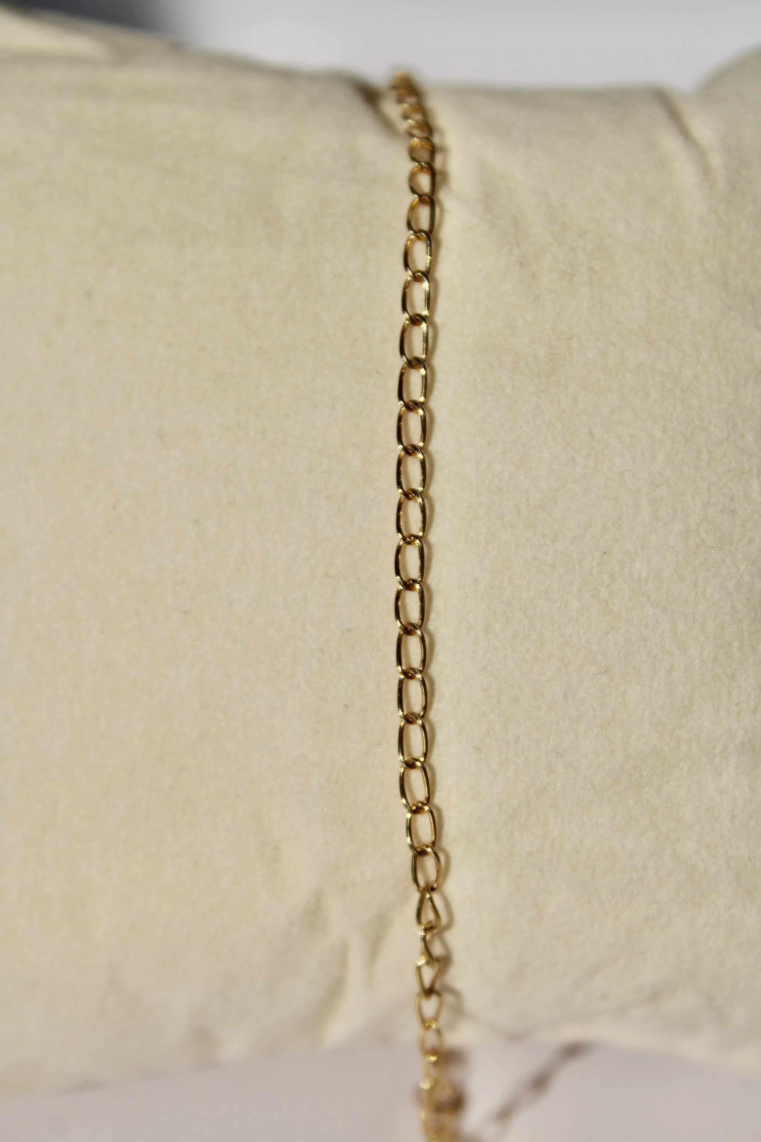 A close-up of a delicate gold chain necklace hanging vertically in front of a beige fabric background.