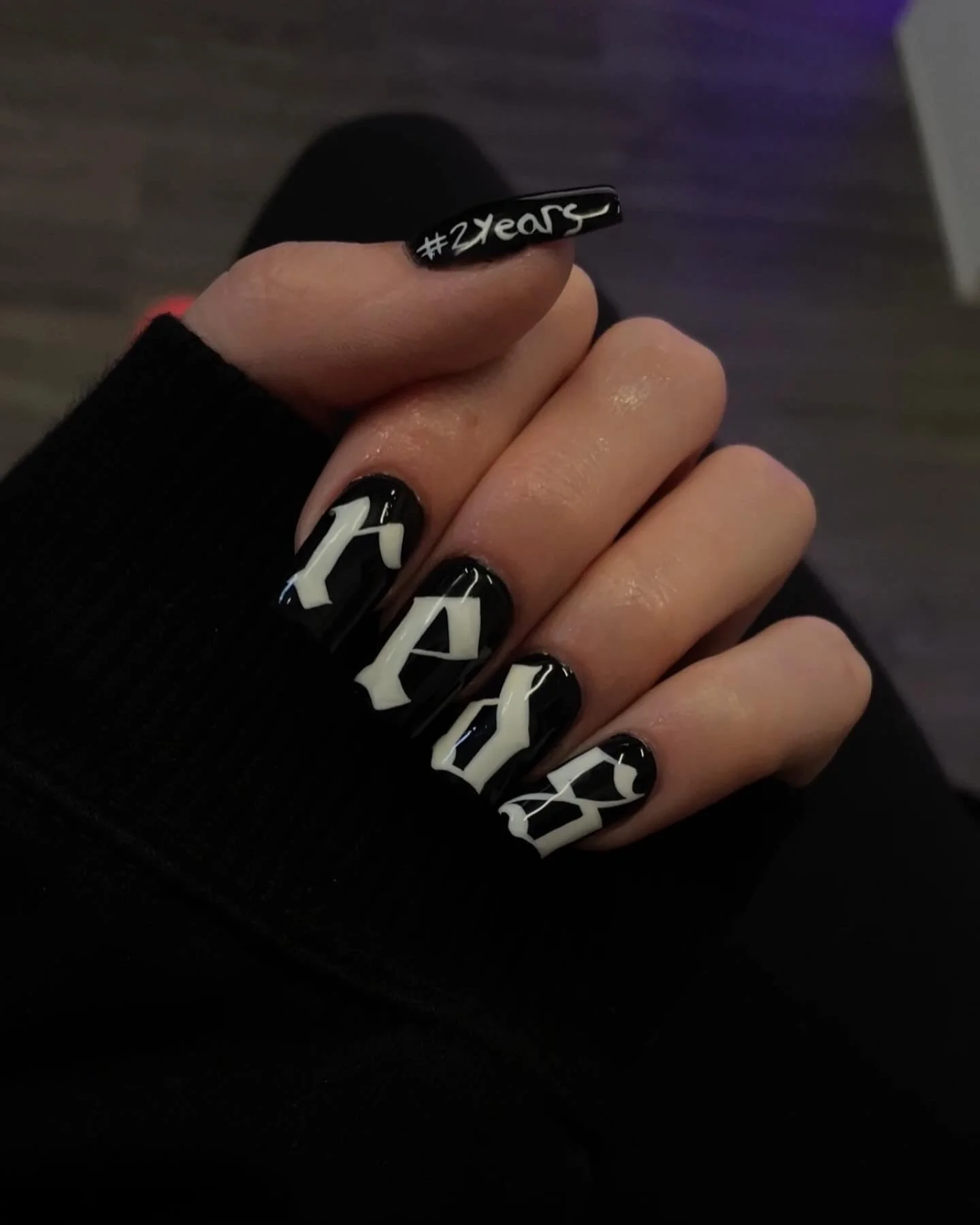 Close-up of a hand with black nail polish and white geometric designs on nails, wearing a black fingerless glove. The thumb's nail is decorated with the text '#2years' in white, on a black background.