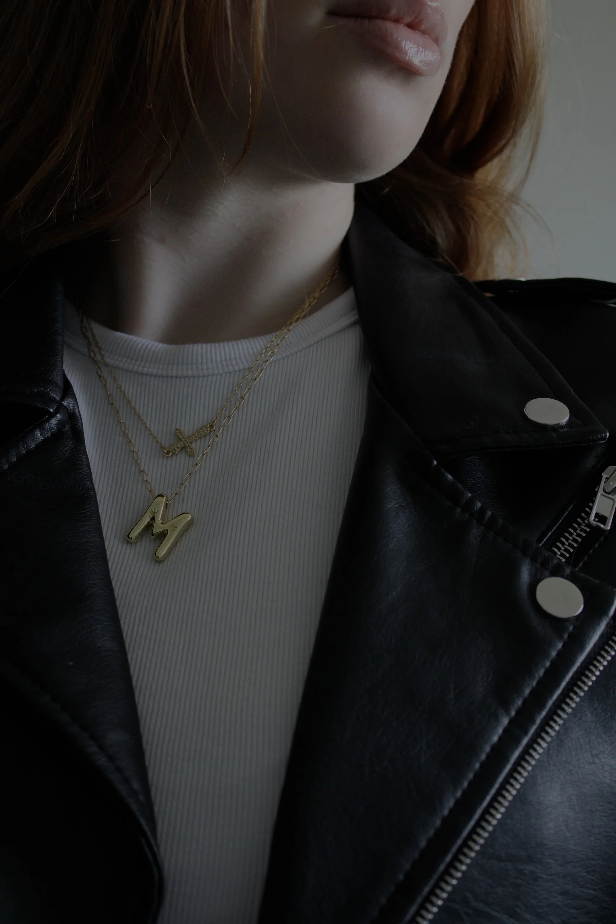 A close-up of a person wearing a black leather jacket, white ribbed shirt, and layered gold necklaces, including one with a handwritten style initial. The person's hair is reddish-brown, and part of their lips and chin are visible.
