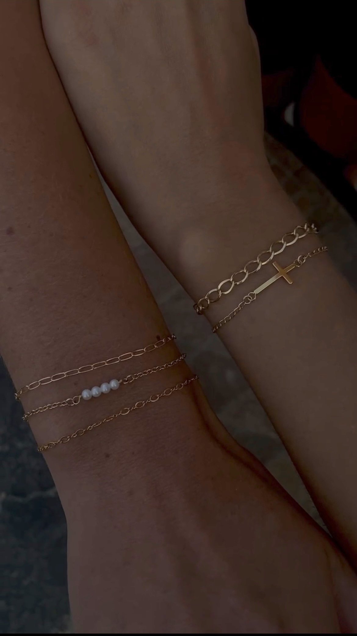 Close-up of three arms with layered gold and pearl bracelets, one with a cross charm, on a person's skin.
