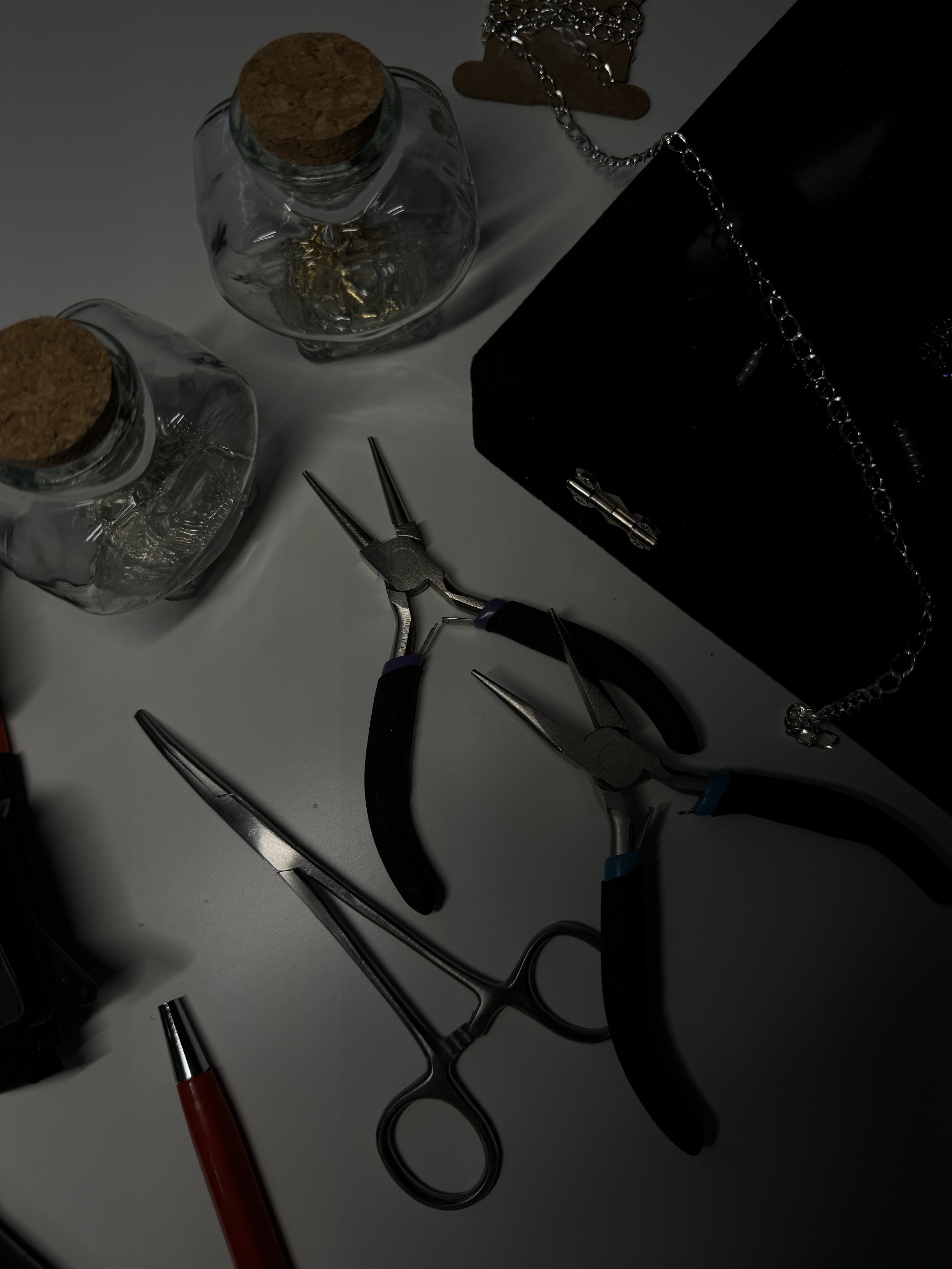 A collection of jewelry-making tools and supplies on a gray surface, including pliers, a pen, glass jars with cork lids, a black box with a chain, a small chain, and a wooden-shaped keychain.