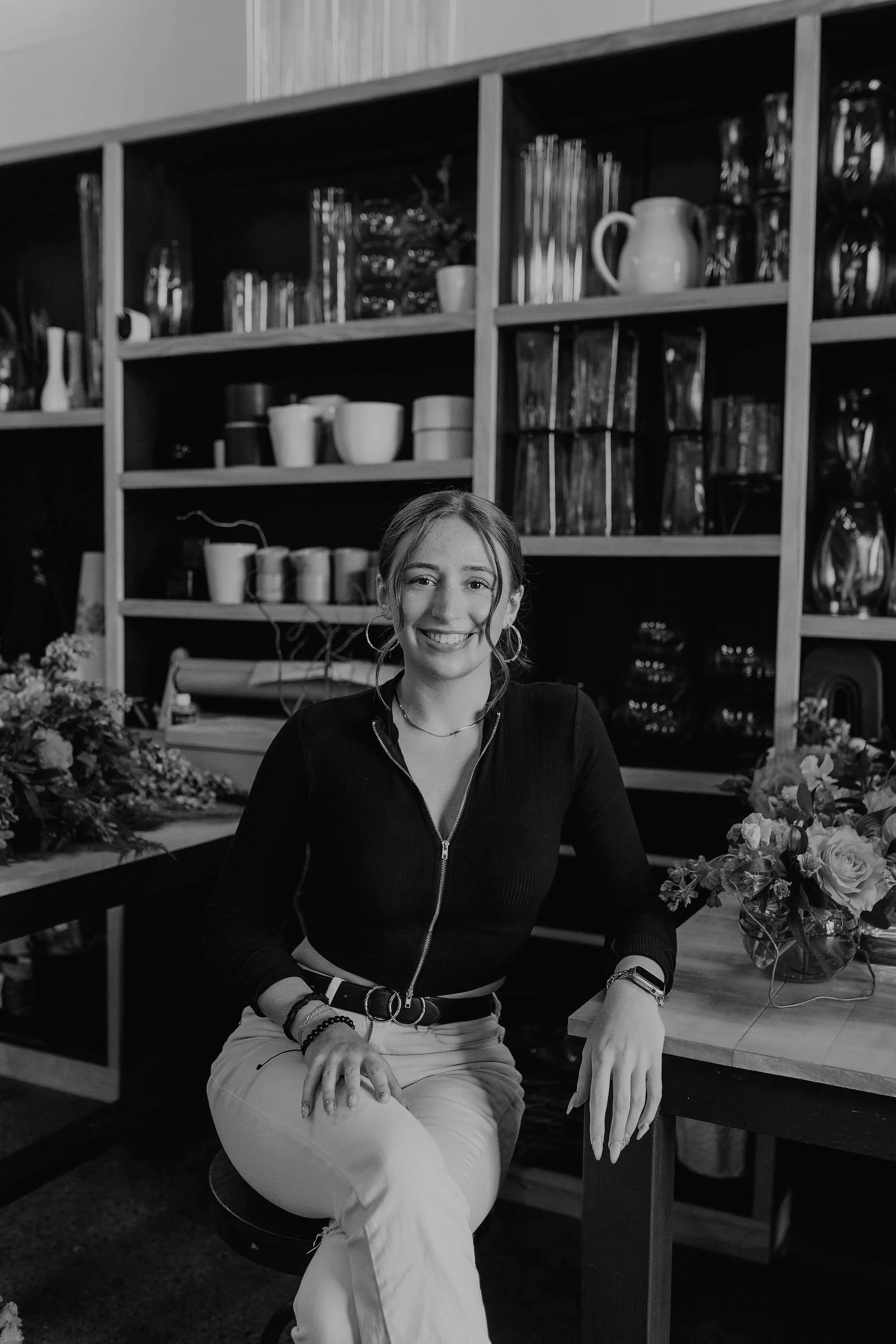 A smiling woman with hoop earrings and bracelets, wearing a zip-up jacket and pants, sitting at a flower shop surrounded by flowers and bouquets, with shelves of glassware and pottery behind her.