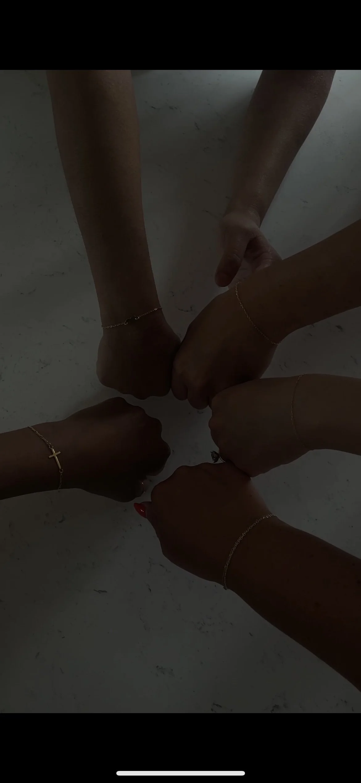 Multiple hands forming a group hug, each hand wearing a delicate gold bracelet or ring, on a light-colored marble surface.