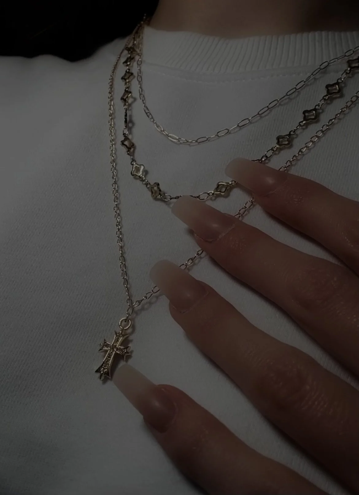Close-up of a person's hand with long, manicured nails, wearing layered silver necklaces with a cross pendant, against a white shirt.
