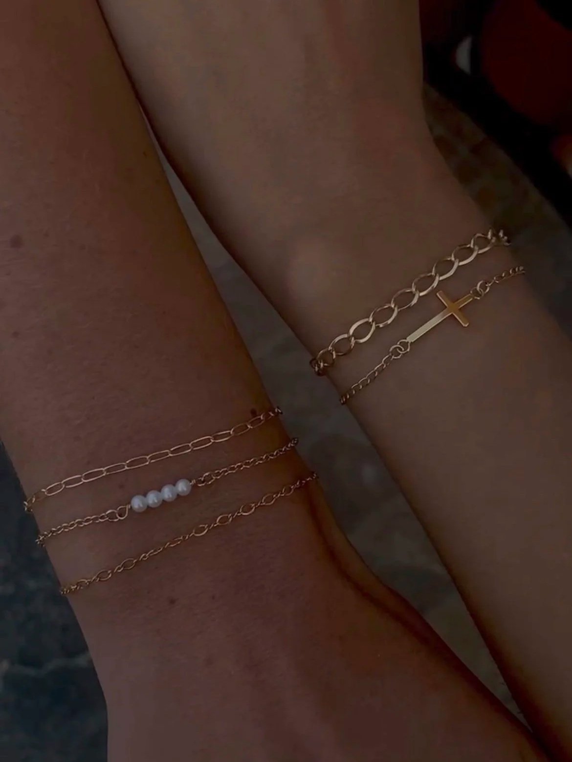 Close-up of a person's wrist and ankle adorned with delicate gold chain bracelets, including one with a cross pendant and another with white beads.