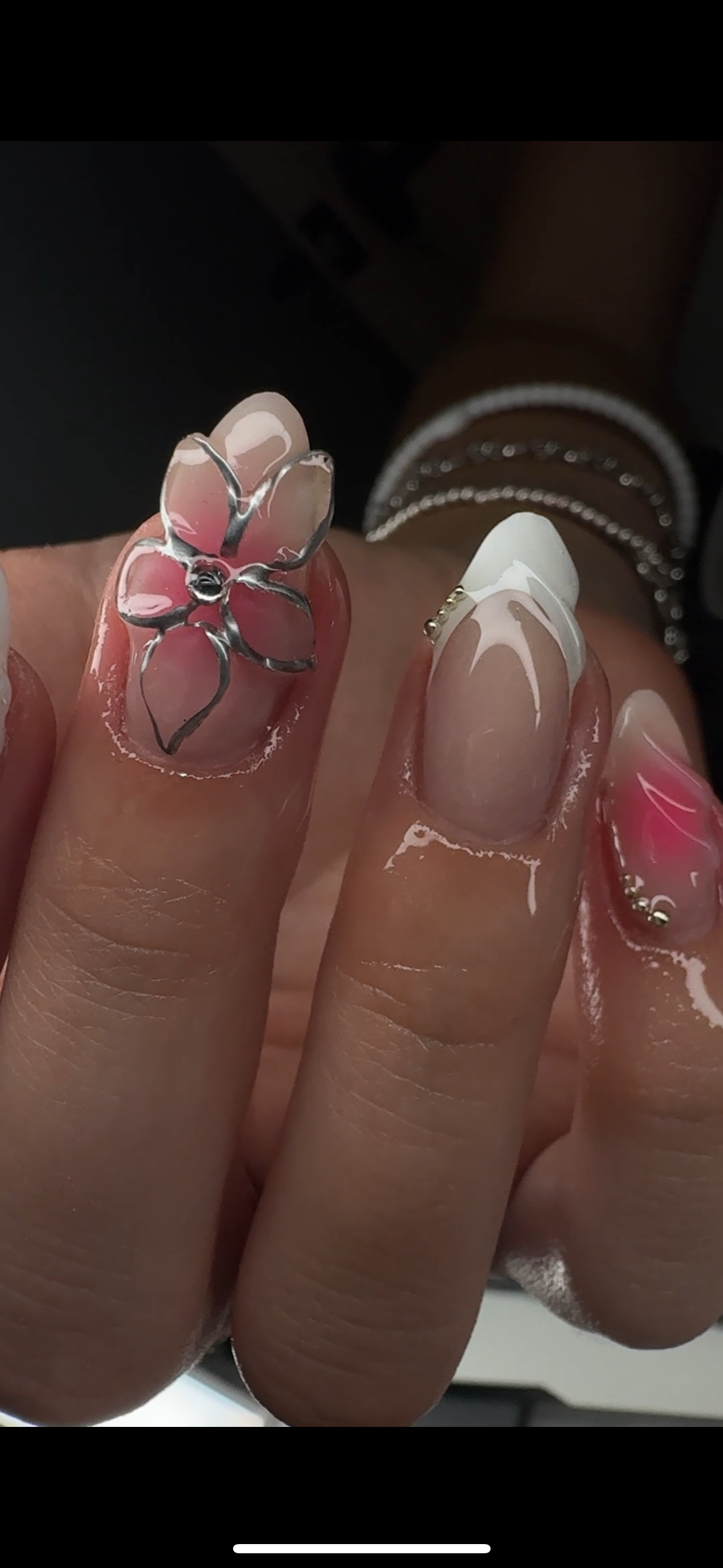 Close-up of a hand with decorated fingernails featuring a pink floral design with silver outlines and rhinestones, and a classic French manicure on another fingernail.
