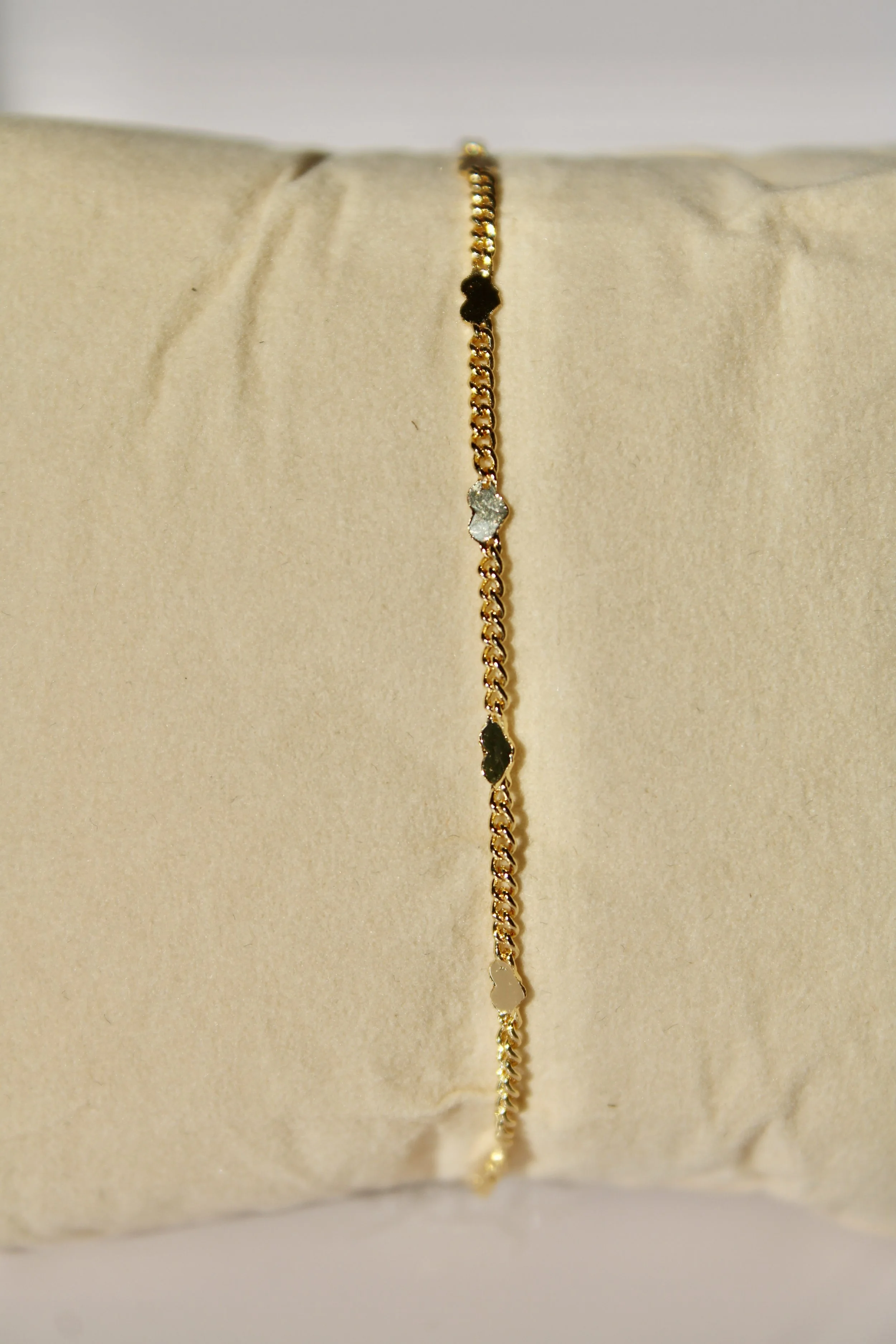 Close-up of a delicate gold chain bracelet with small, evenly spaced charms, displayed on a beige fabric pillow.