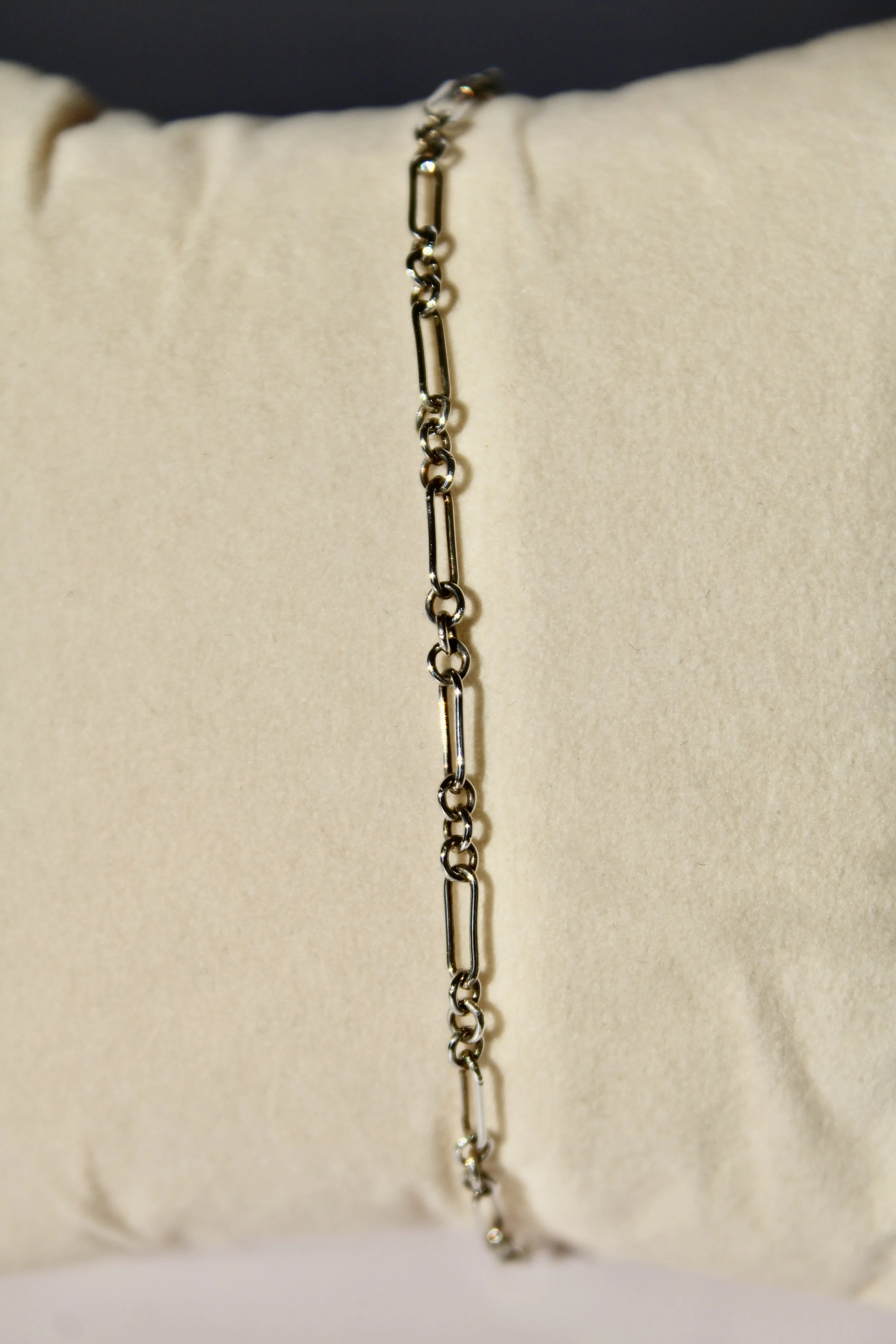Close-up of a silver chain bracelet draped over a rolled white fabric display. The chain features elongated rectangular links and smaller circular links.