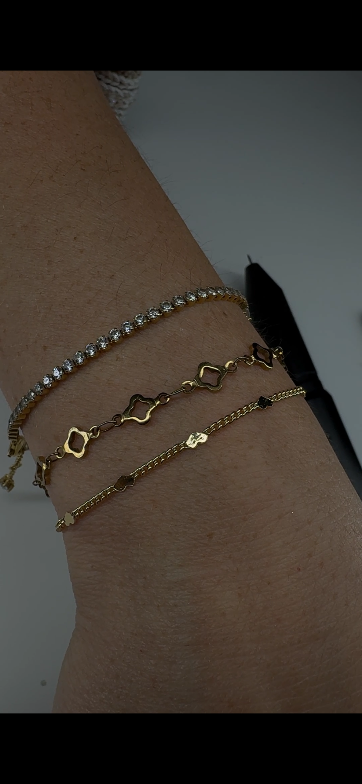 Close-up of four gold bracelets on a person's wrist. The top bracelet has clear gemstones, the second features geometric shapes, the third is a simple chain with small black details, and the bottom is a delicate chain with a small charm.