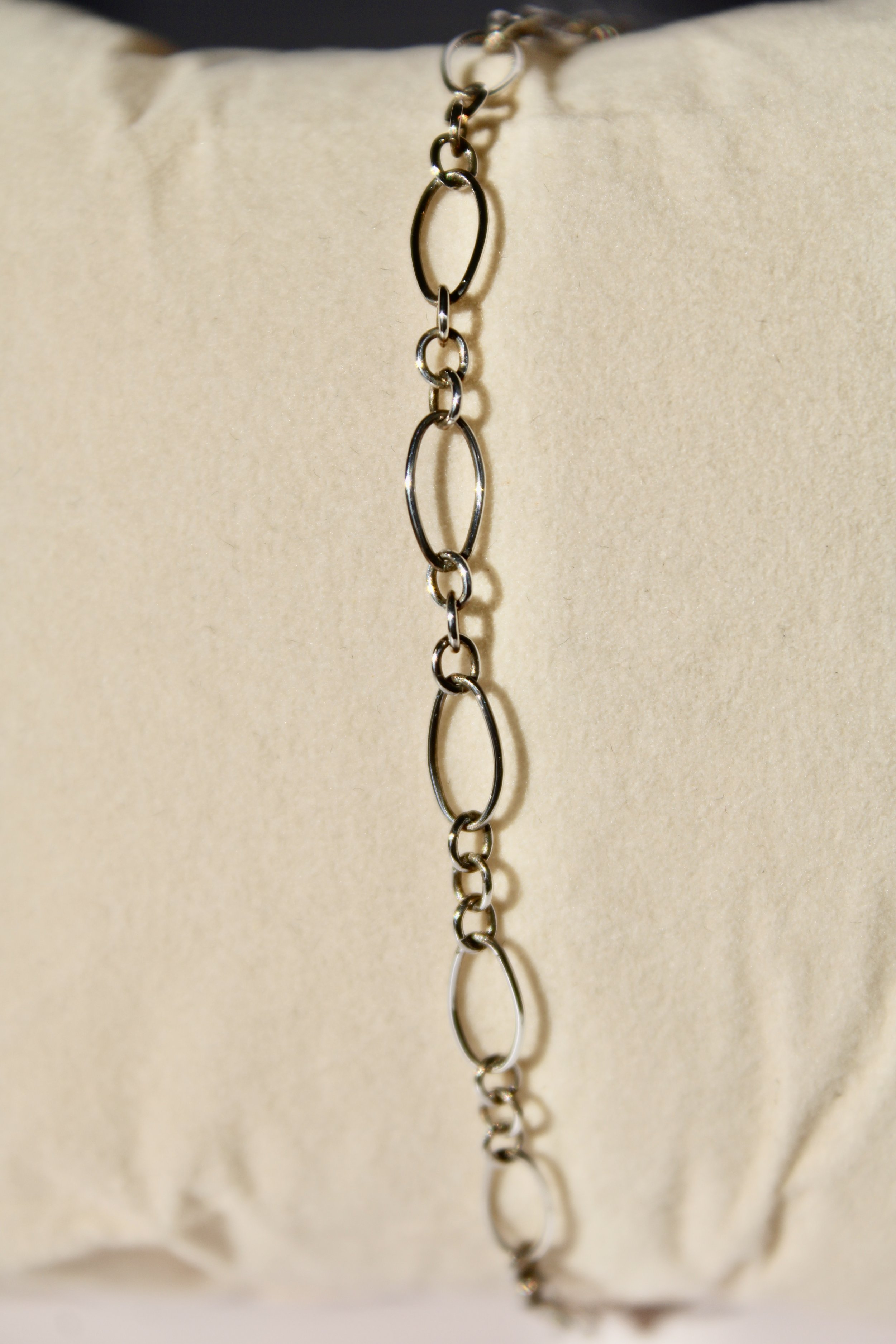 Close-up of a silver chain necklace with various oval and circular links, hanging against a soft beige background.