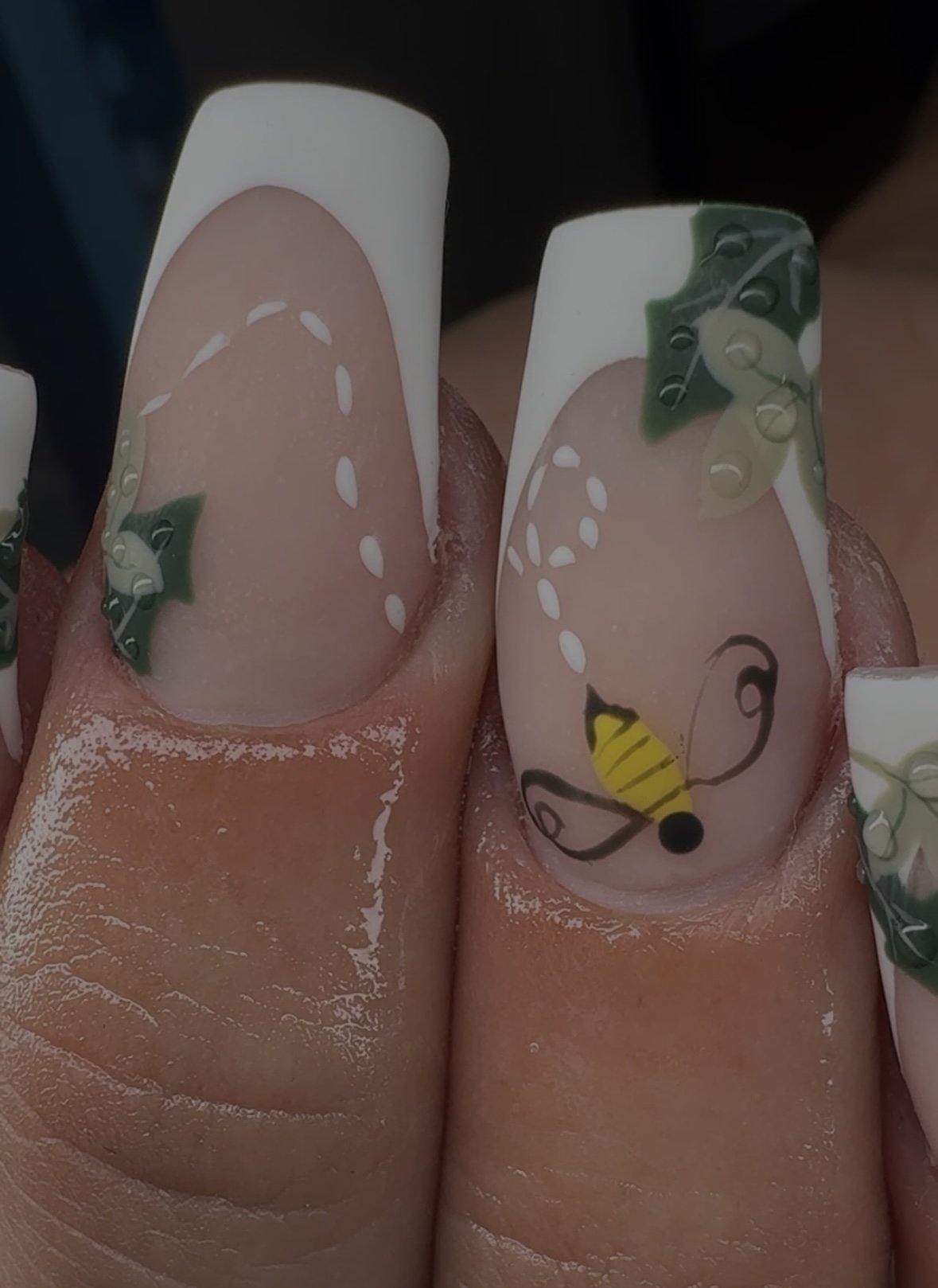Nail art on two fingers depicting Christmas themes, with holly leaves, a yellow and black striped bee, and white dashed lines on a pinkish background.