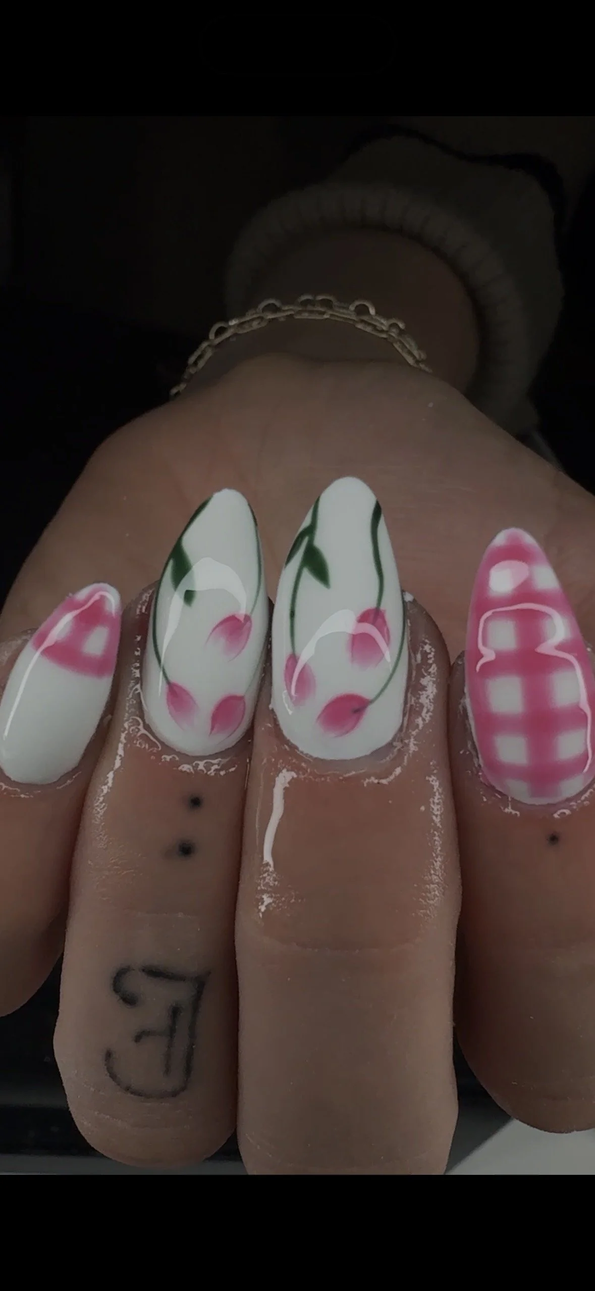 Close-up of four fingers with decorated gel nails: the first finger has a pink, white, and green cherry design; the second and third fingers feature a white base with pink cherry blossom flowers and green stems; the fourth finger has a pink and white checkered pattern. The fingers have tattoos and are positioned against a dark background.